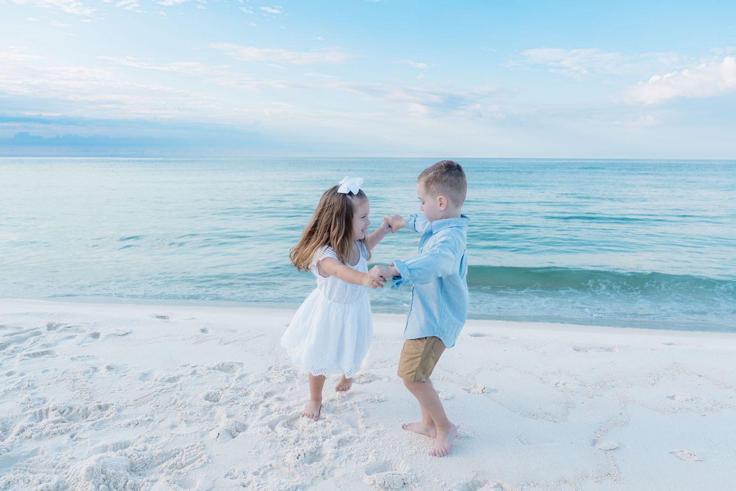 Two young children in white and blue outfits hold hands and play on a white sandy beach with calm turquoise water.