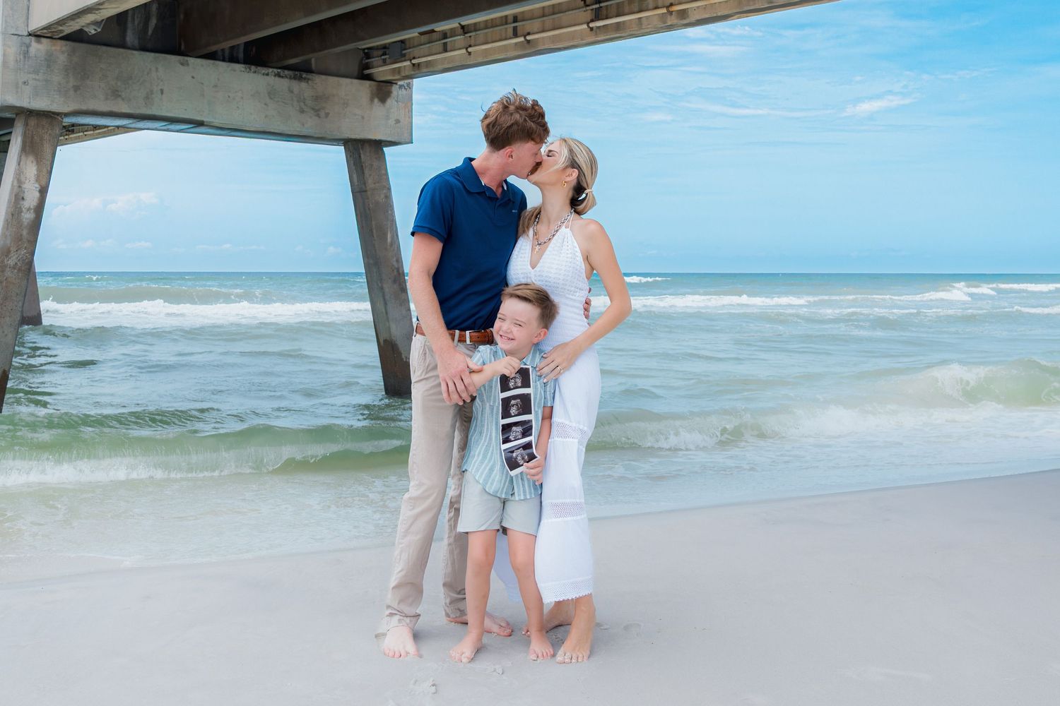 Family of three poses on beach near pier, couple kissing as child holds ultrasound photos announcing pregnancy.