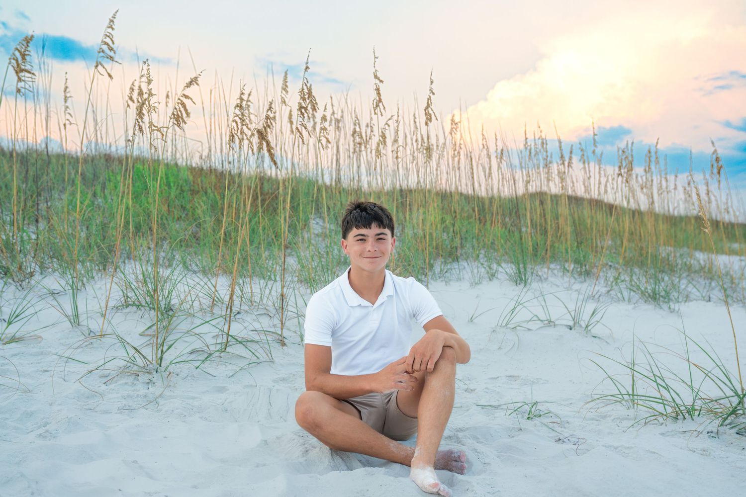 Teen boy in white polo sits on white sand dunes with sea oats at sunset on a Florida beach.