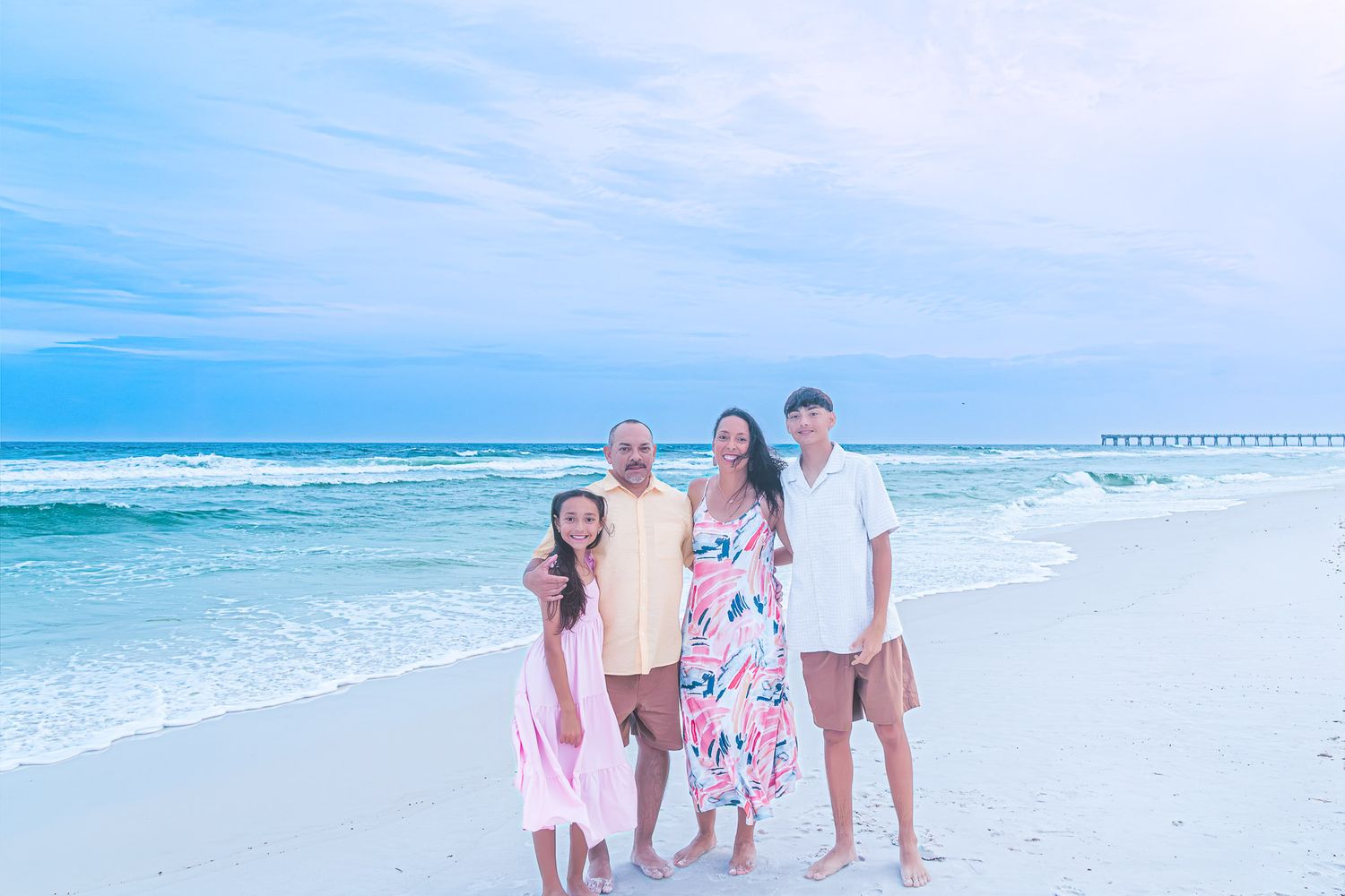 Family of four smiles together on white sandy Florida beach with pier and blue ocean in background.