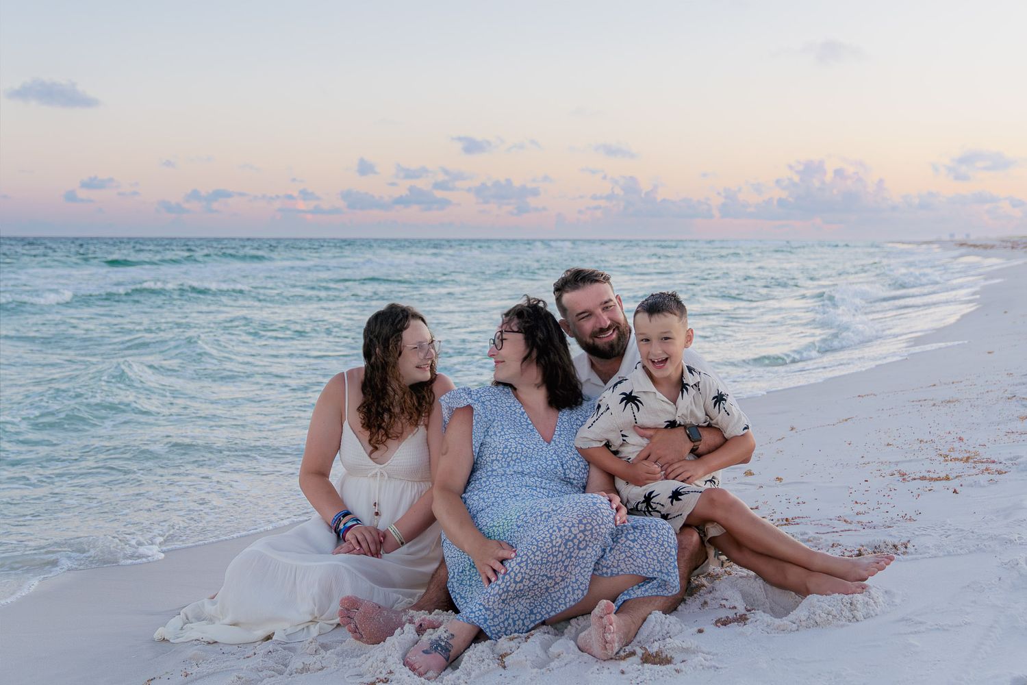 Family of four sitting together on a sandy beach at sunset, smiling and laughing with calm turquoise ocean waves behind them.
