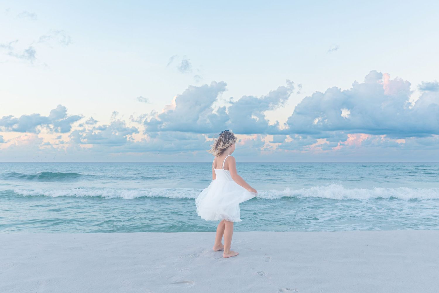 A young girl in a white dress twirls on a sandy beach with turquoise waves and pastel clouds behind her.