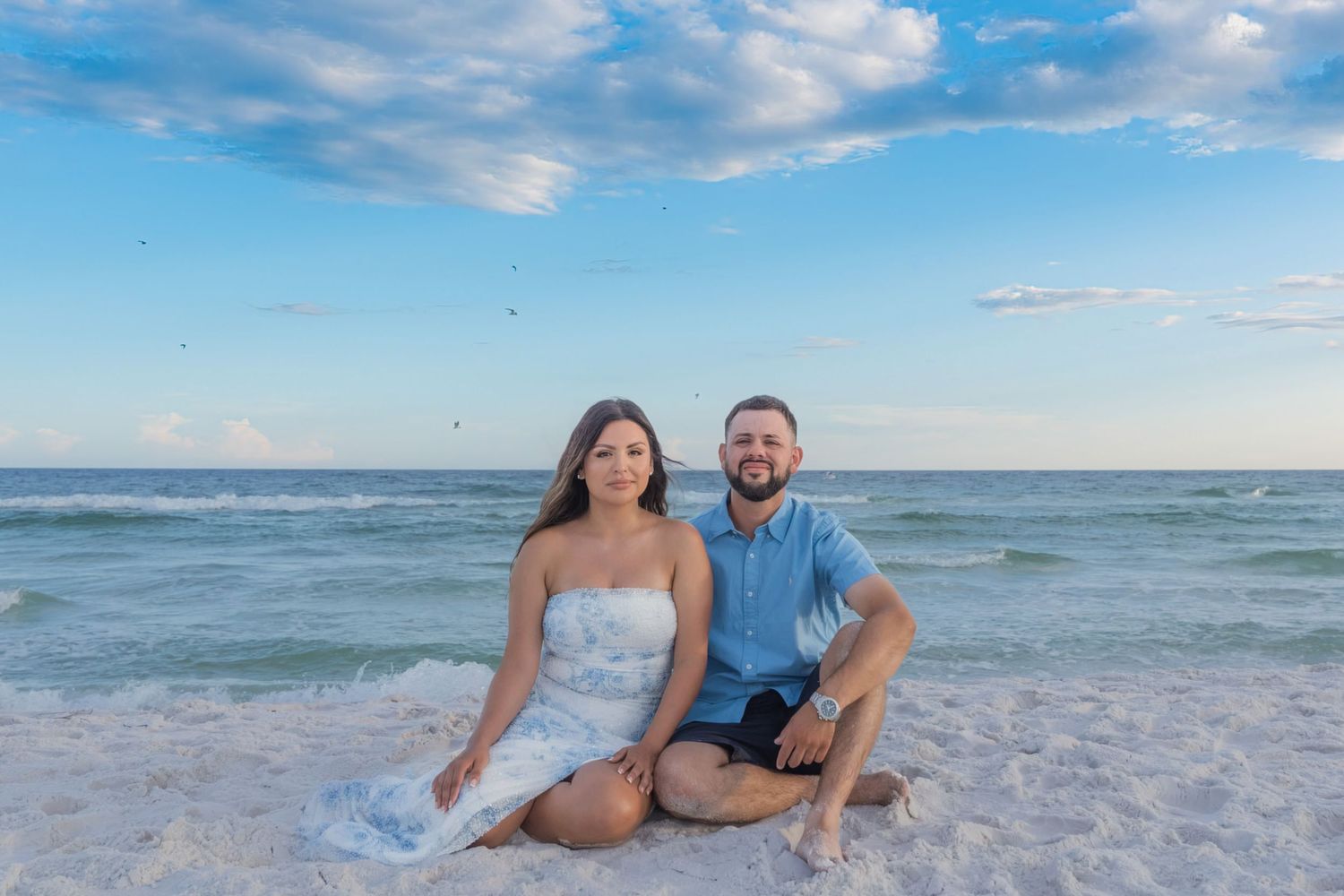 Couple sitting together on white sand beach with turquoise ocean waves and blue sky backdrop.