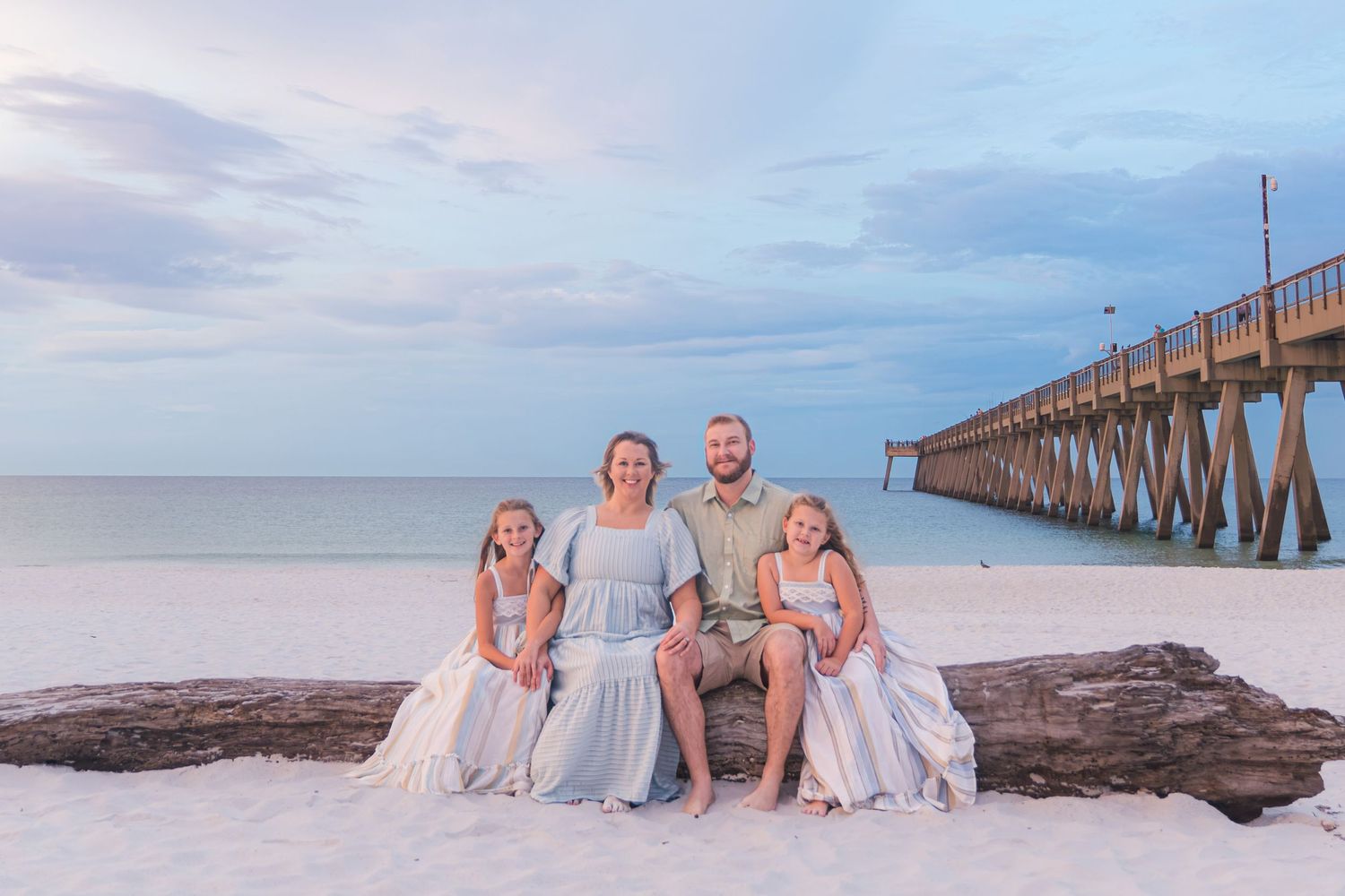 Family of four poses on driftwood at a white sand beach near a pier at sunset.