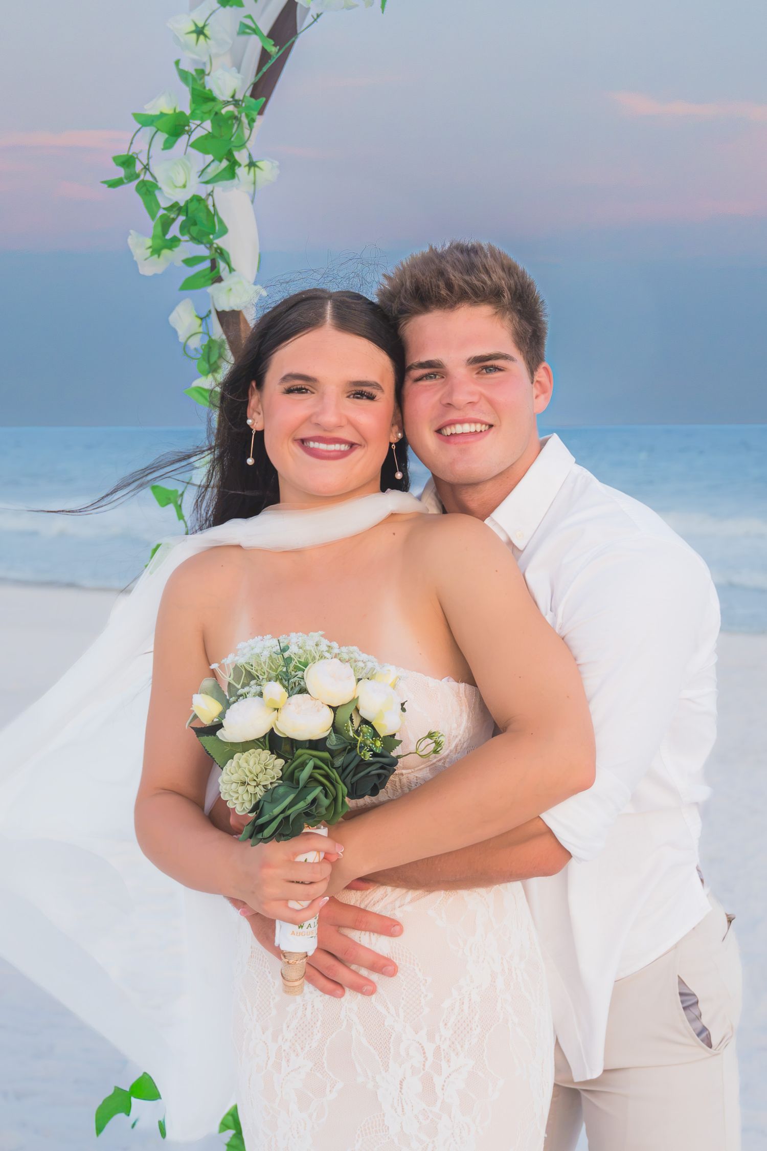 Smiling bride and groom embrace under floral arch on beach at sunset, bride holding white tulip bouquet.
