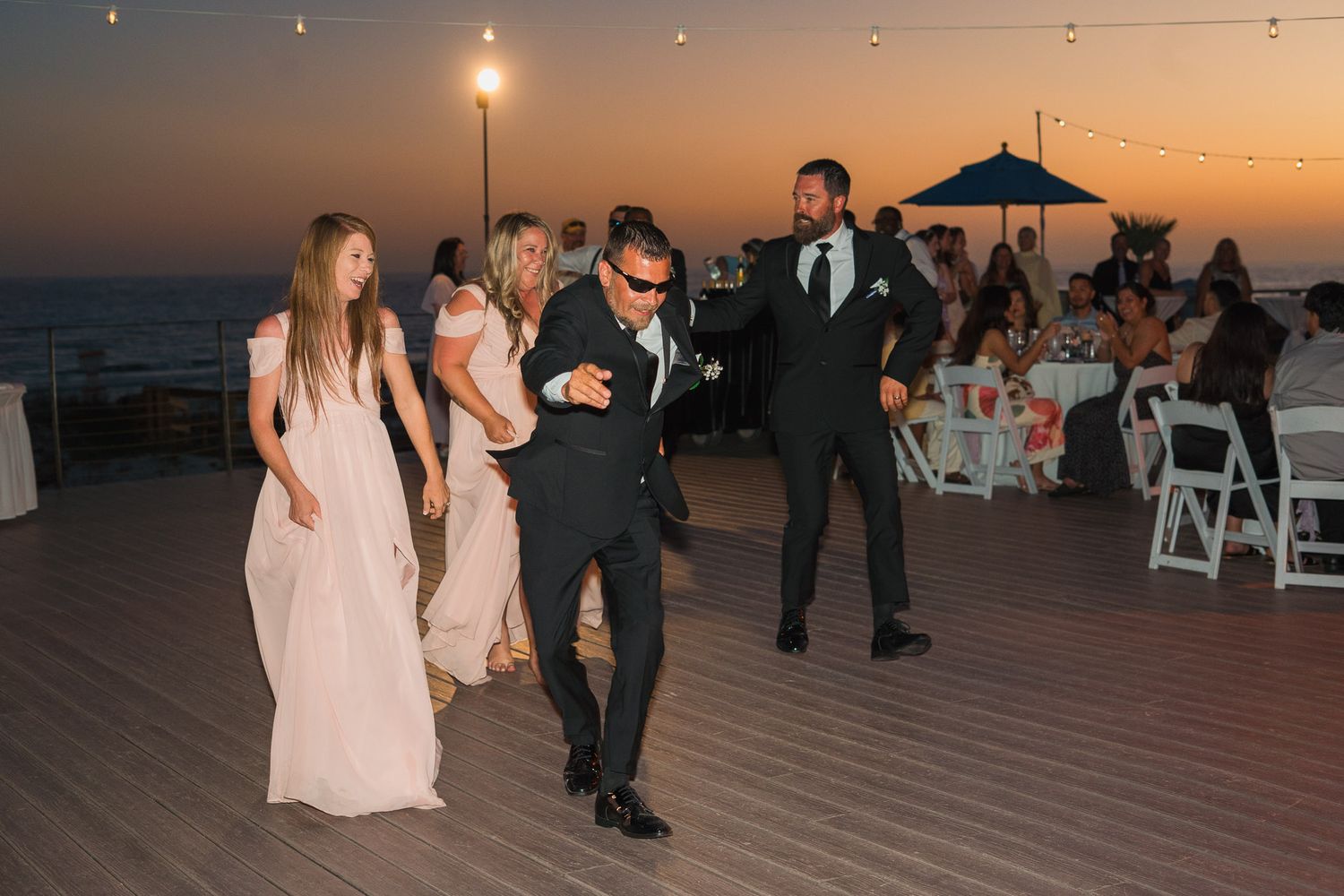 Guests dancing on an outdoor deck at a wedding reception during a stunning sunset over the ocean.