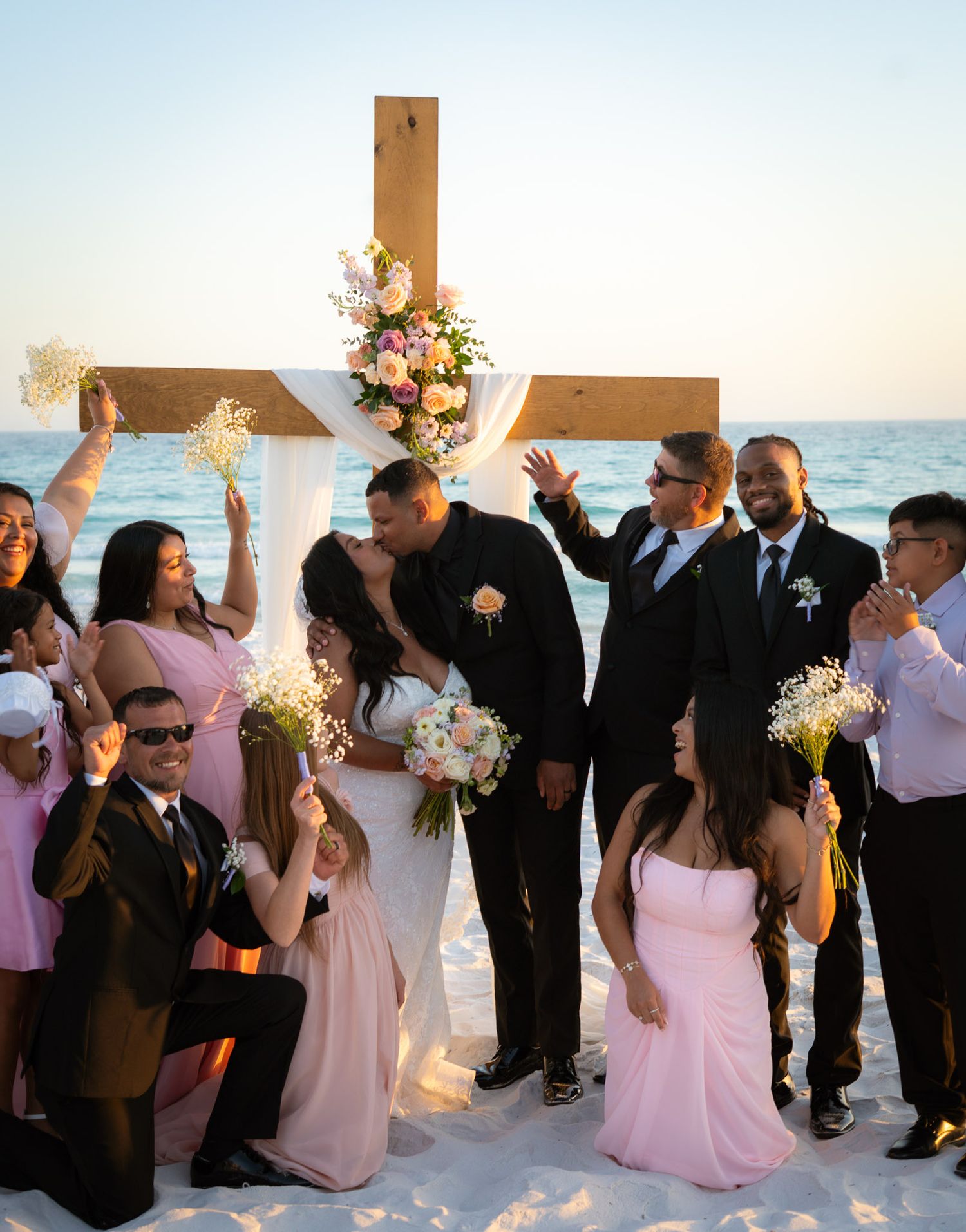 Wedding party celebrates with the bride and groom kissing before a wooden cross altar on a sunny beach.