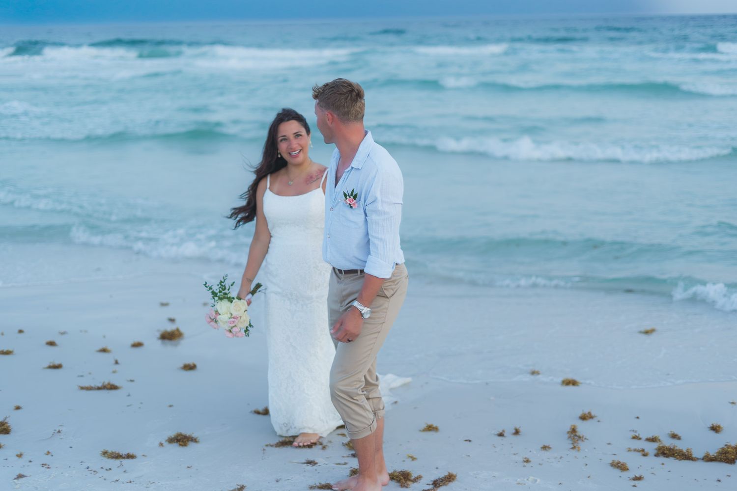 Couple in wedding attire embracing on a white sand beach with turquoise ocean waves in the background.