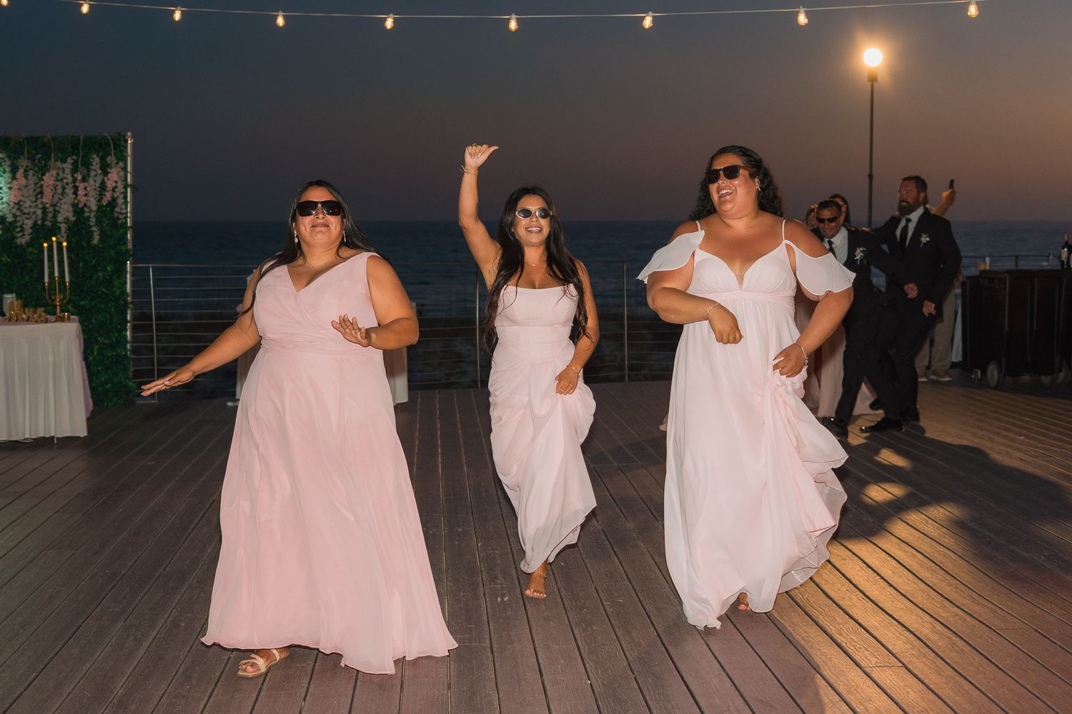Three bridesmaids in blush pink gowns dancing joyfully on an outdoor deck at a sunset wedding reception.