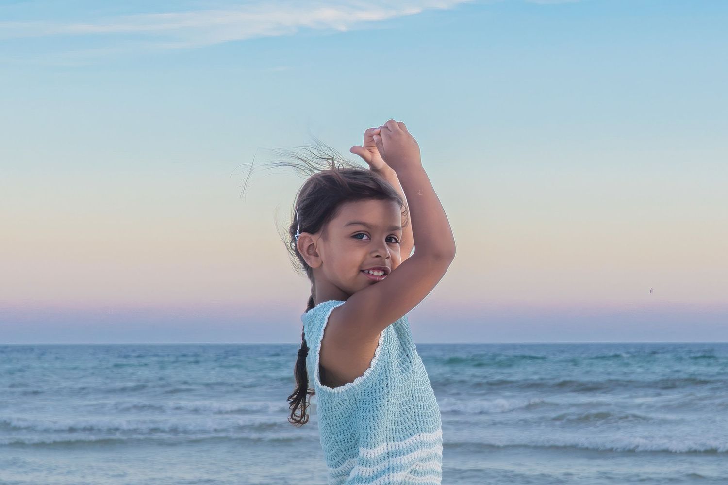 Young girl with braids poses on beach at sunset, arms raised, wearing light blue sleeveless top with ocean backdrop.