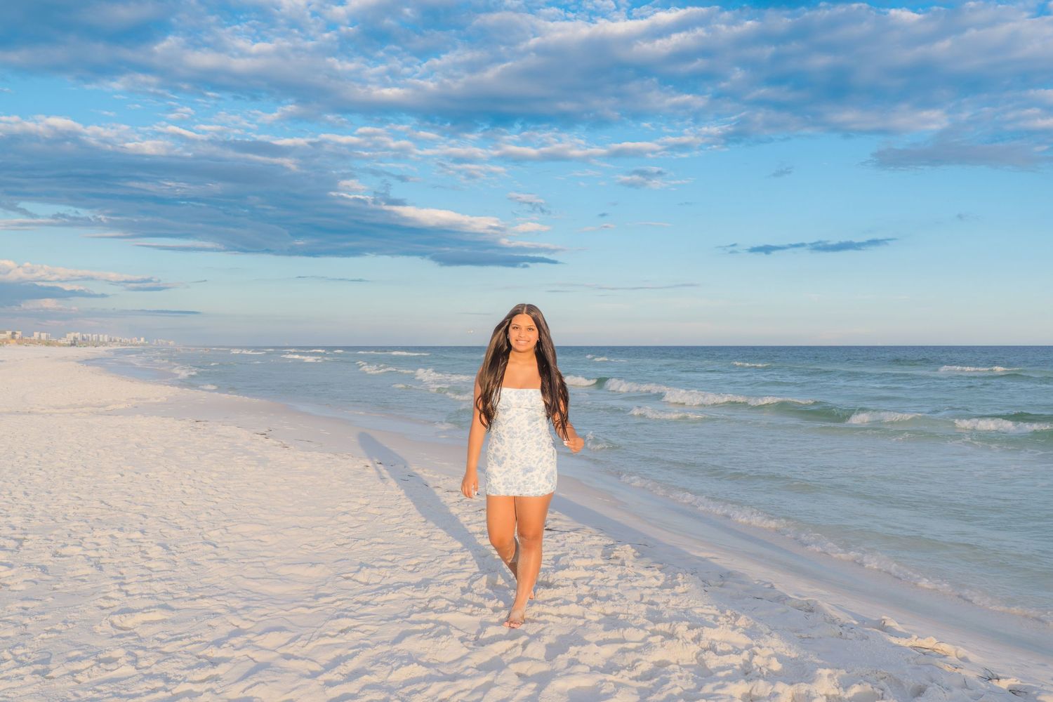 A young woman in a white floral dress walks barefoot on a sunny white sand beach with blue skies and ocean waves.