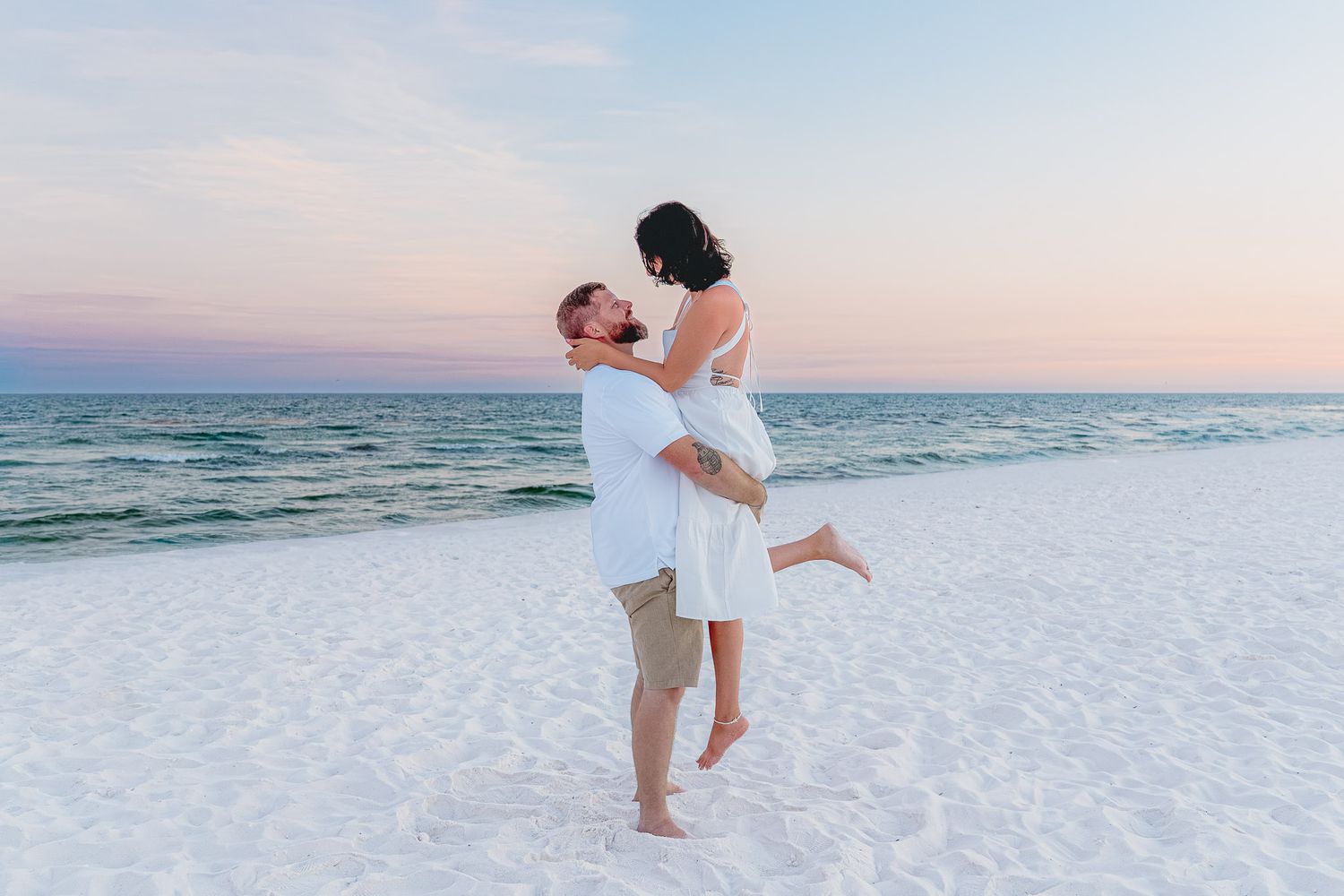 Man lifting woman in white dress on sandy beach during pink sunset by the ocean.