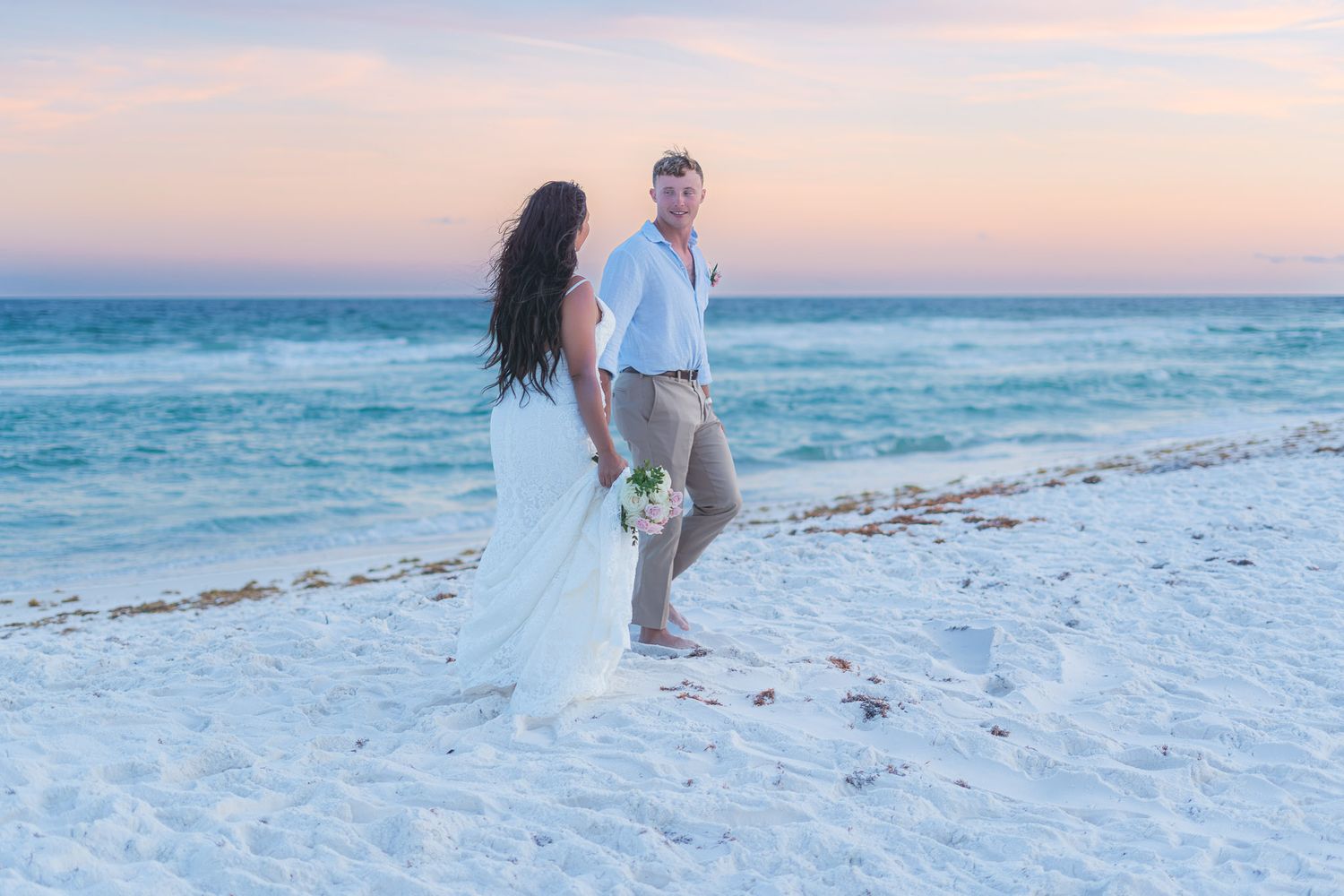 Bride and groom hold hands on a white sand beach at sunset during their romantic destination wedding ceremony.