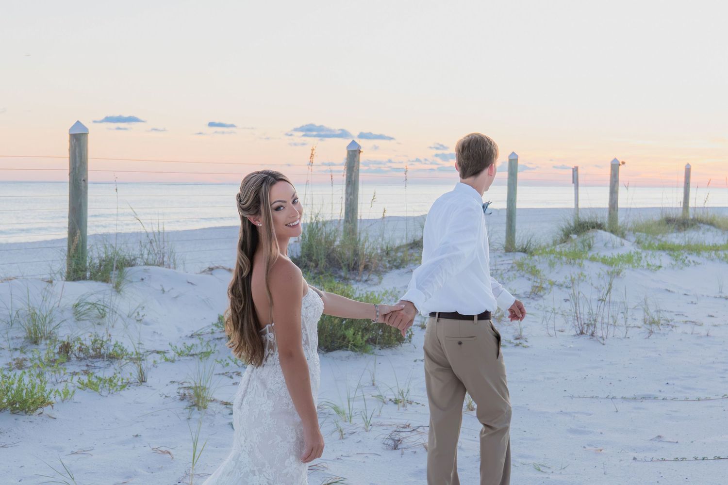 Bride and groom hold hands on sandy beach at sunset, bride in lace gown with flowing hair at coastal wedding.
