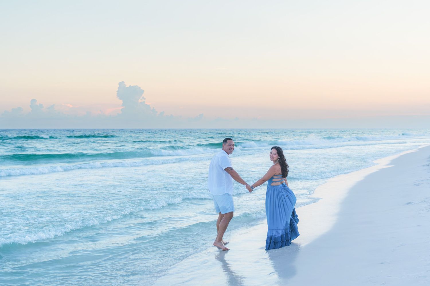 Couple holding hands walking on a white sandy beach at sunset with turquoise ocean waves.