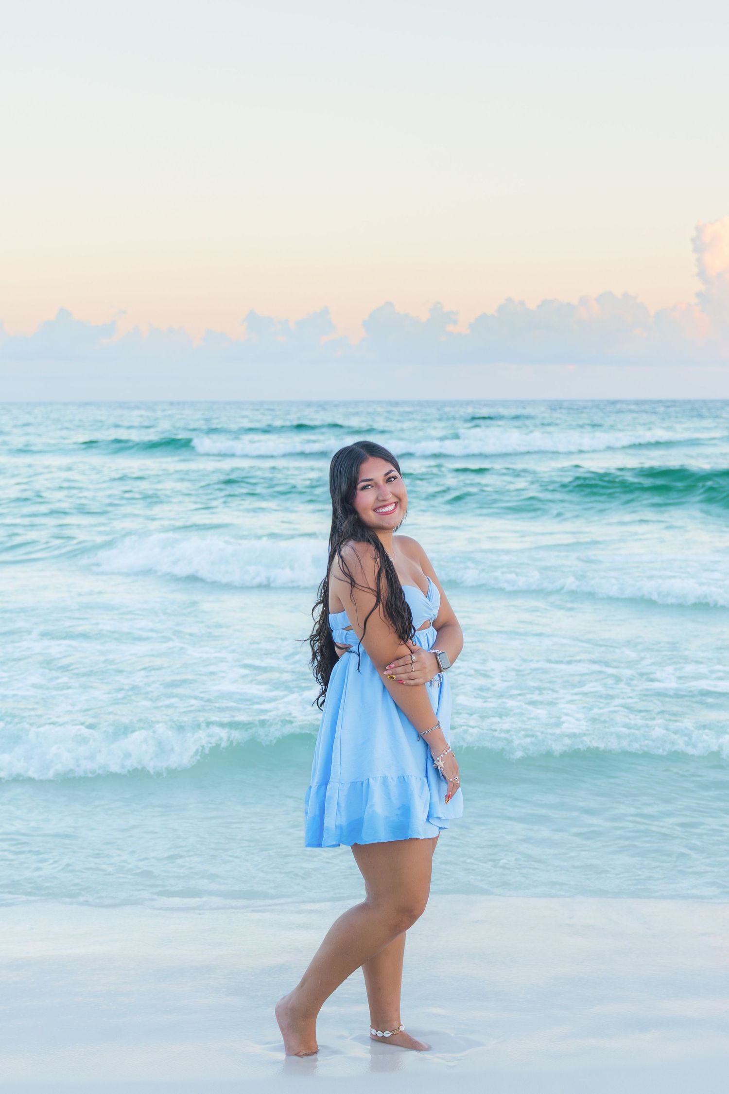 Young woman in light blue dress smiling on a sandy beach with turquoise ocean waves at sunset.