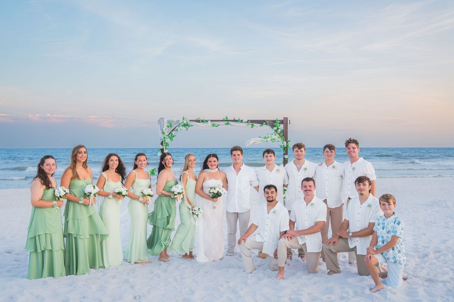 Wedding party poses on sandy beach at sunset with green floral arch backdrop.