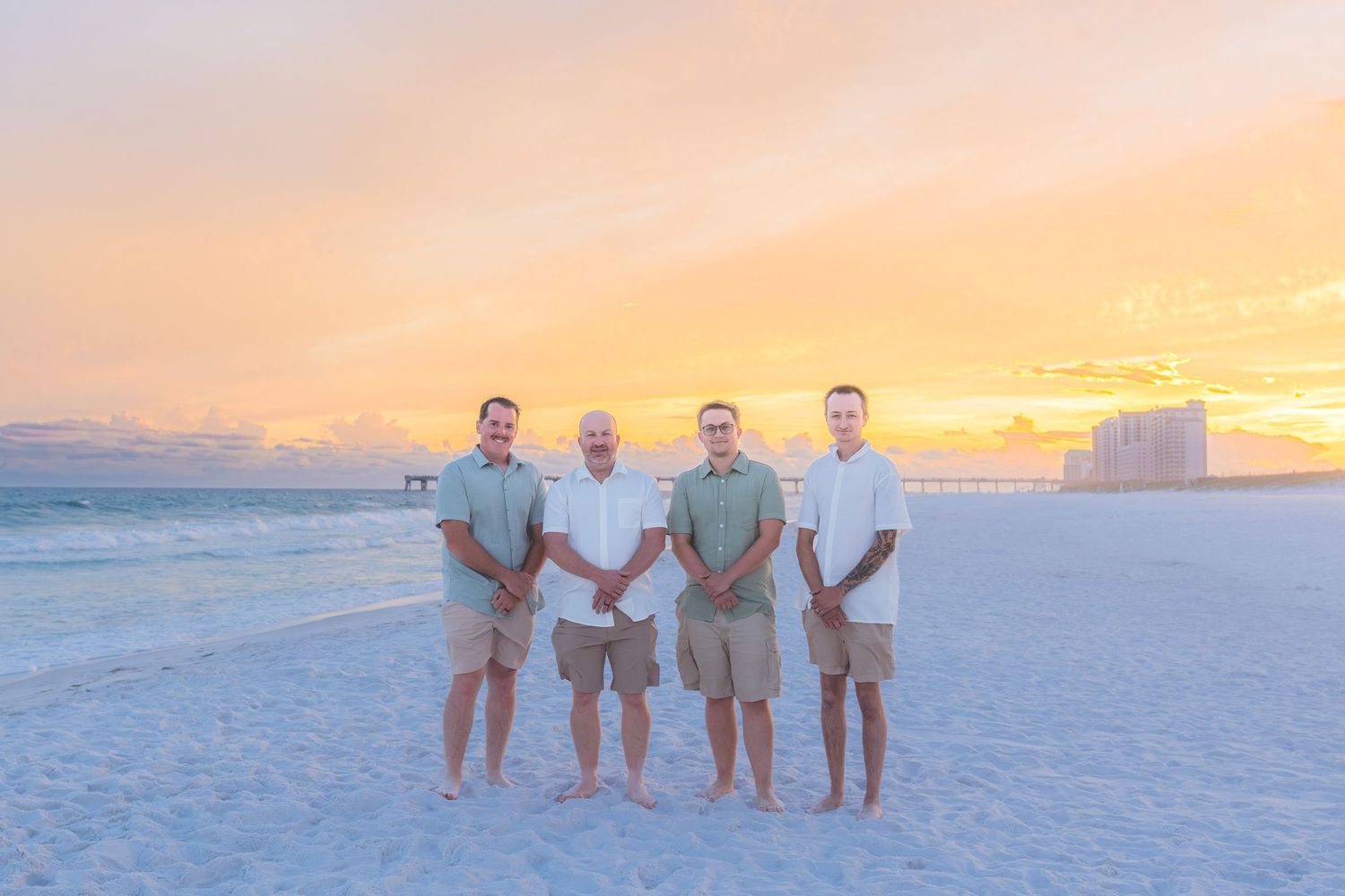 Four men in matching outfits pose on a beach at sunset with a colorful pink and orange sky behind them.