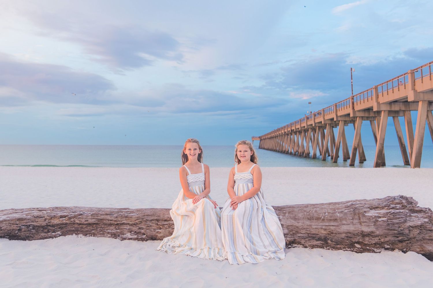 Two girls in white dresses sit on driftwood at a white sand beach with a wooden pier at sunset.