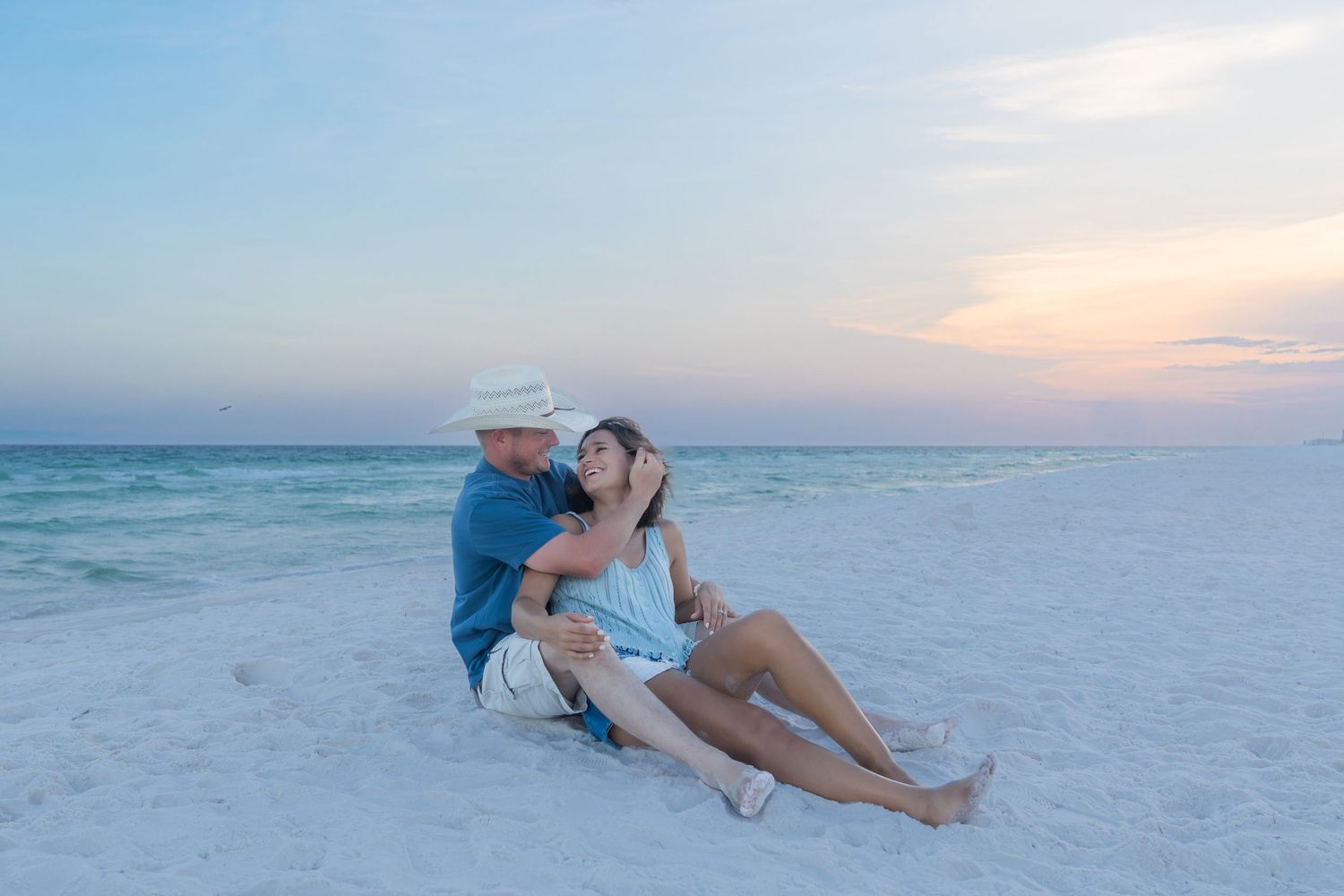 Couple sitting together on white sand beach at sunset, man in hat embracing woman in blue dress by calm turquoise ocean.