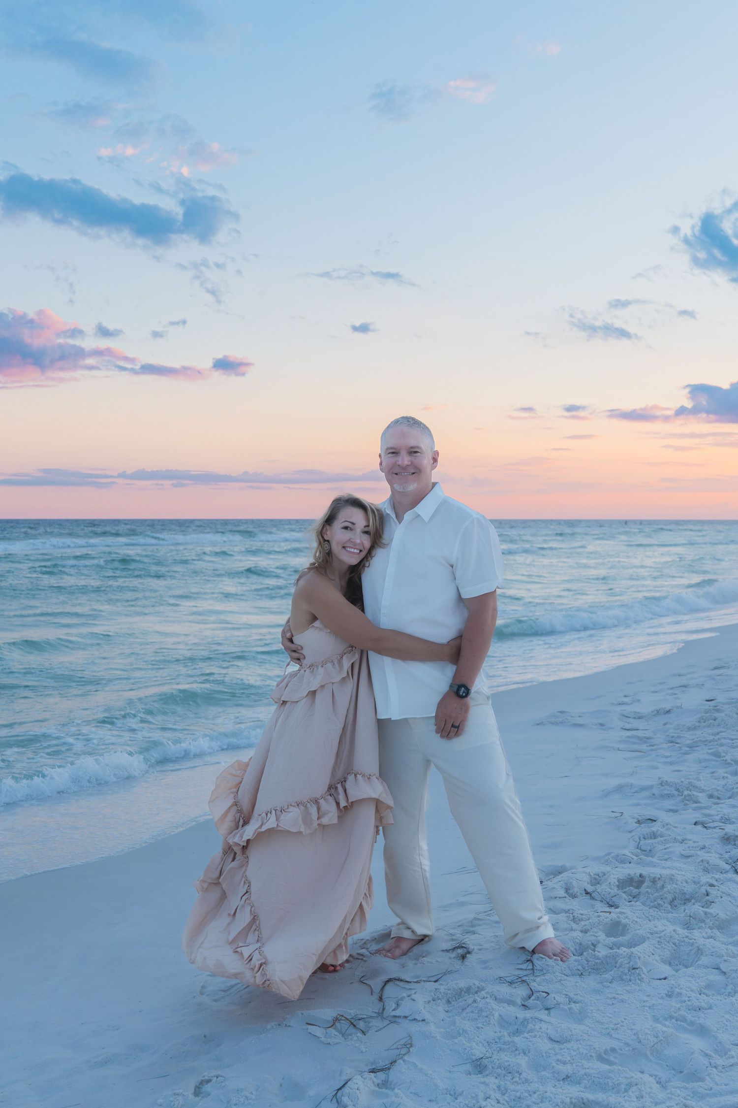 Couple embracing on a sandy beach at sunset, woman in flowing pink dress, man in white shirt and khaki pants.