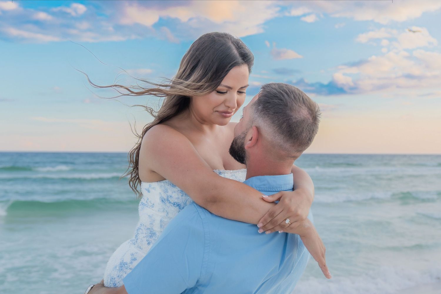 Couple embracing on a beach at sunset, woman in white dress with wind-blown hair, man in light blue shirt.