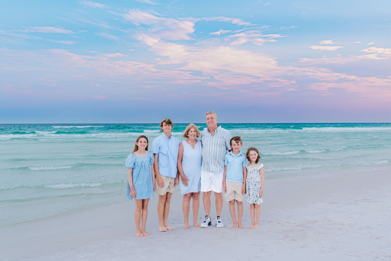 Family of six posing on a white sand beach at sunset with turquoise water and pink sky backdrop.