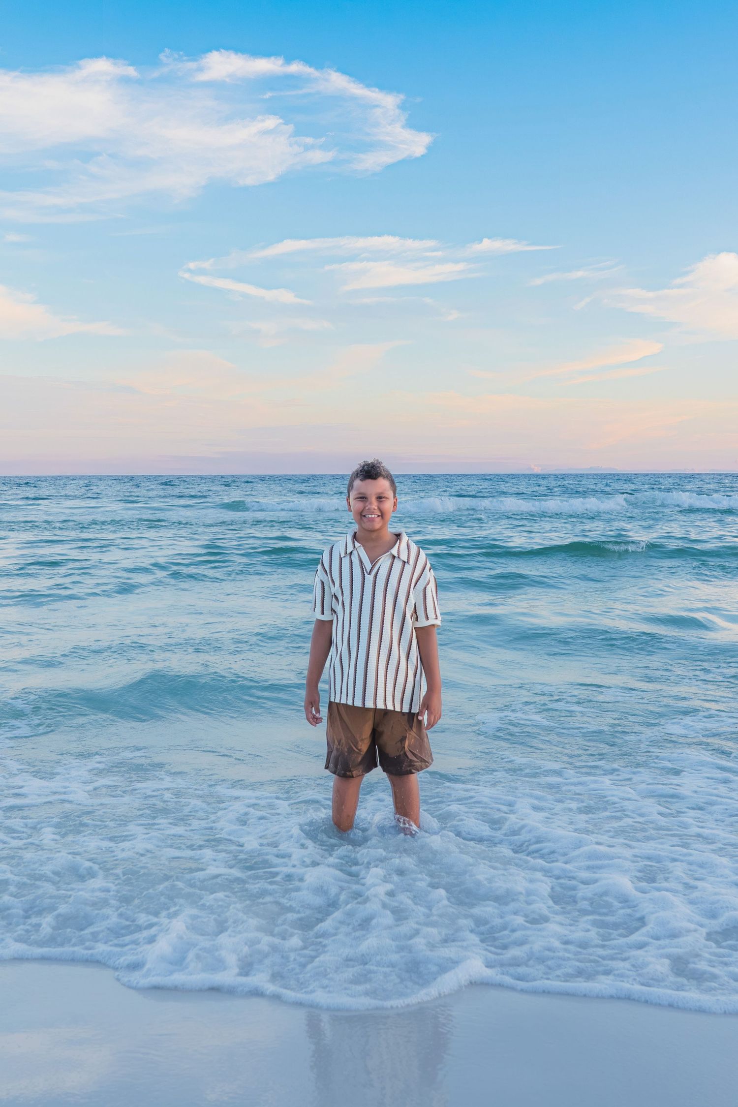 A smiling boy stands in shallow ocean waves wearing a striped shirt and brown shorts on a beach at dusk.