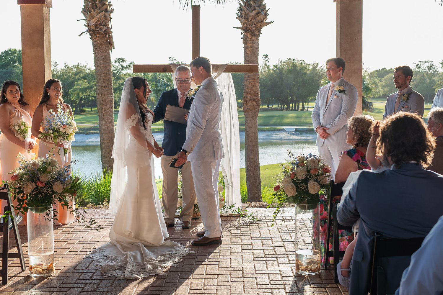 Outdoor wedding ceremony by a lake, couple exchanging vows under a wooden arch with floral arrangements.