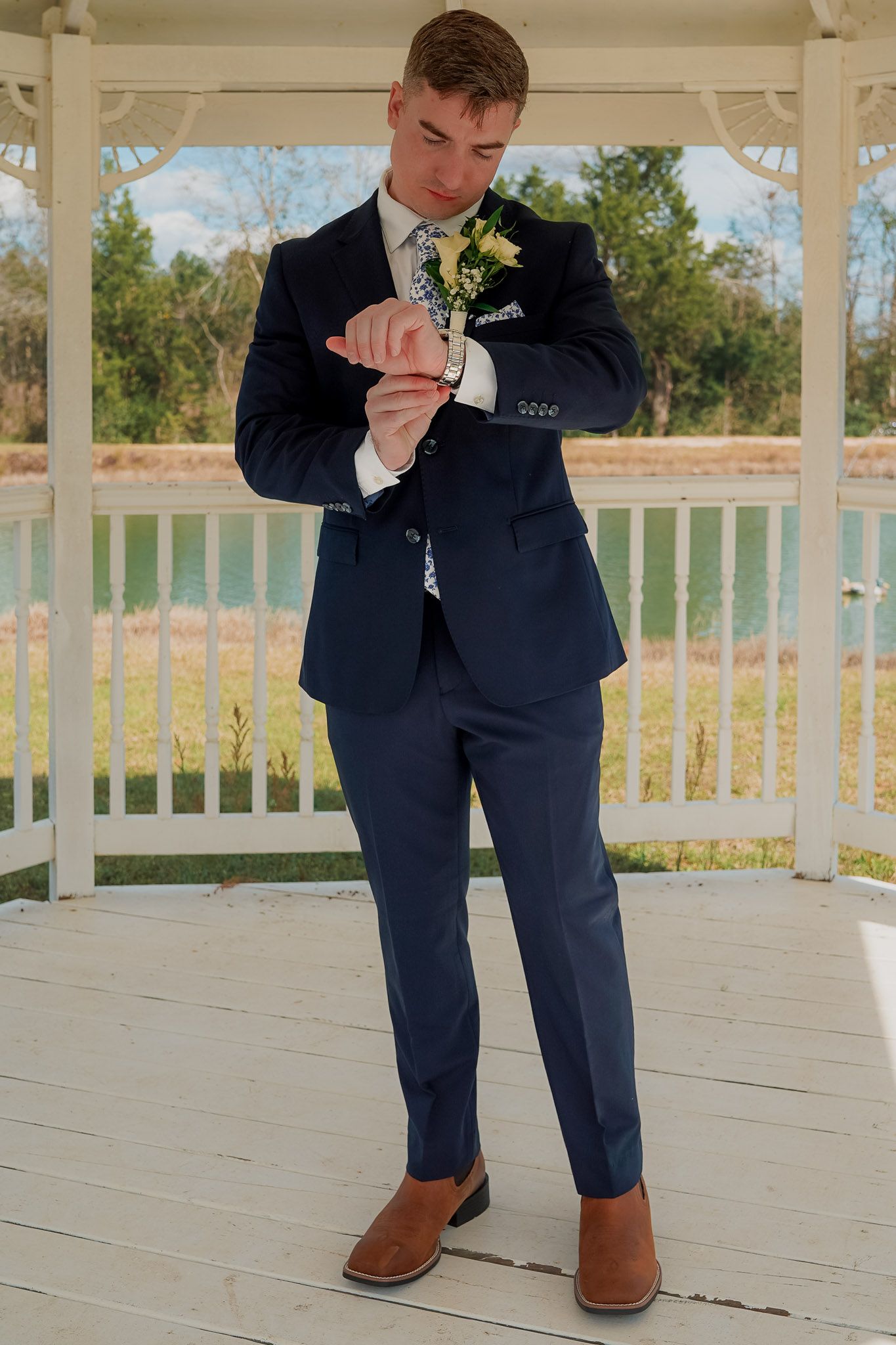 Young man in navy blue suit with boutonniere adjusting cufflinks under white gazebo outdoors.