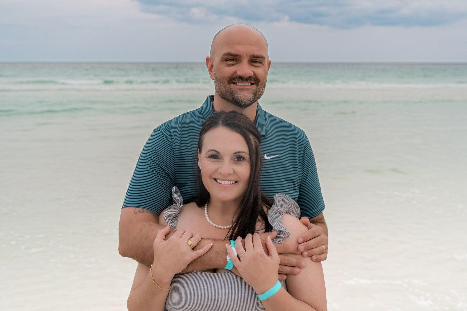 Couple smiling together on a white sand beach with turquoise ocean water in the background.