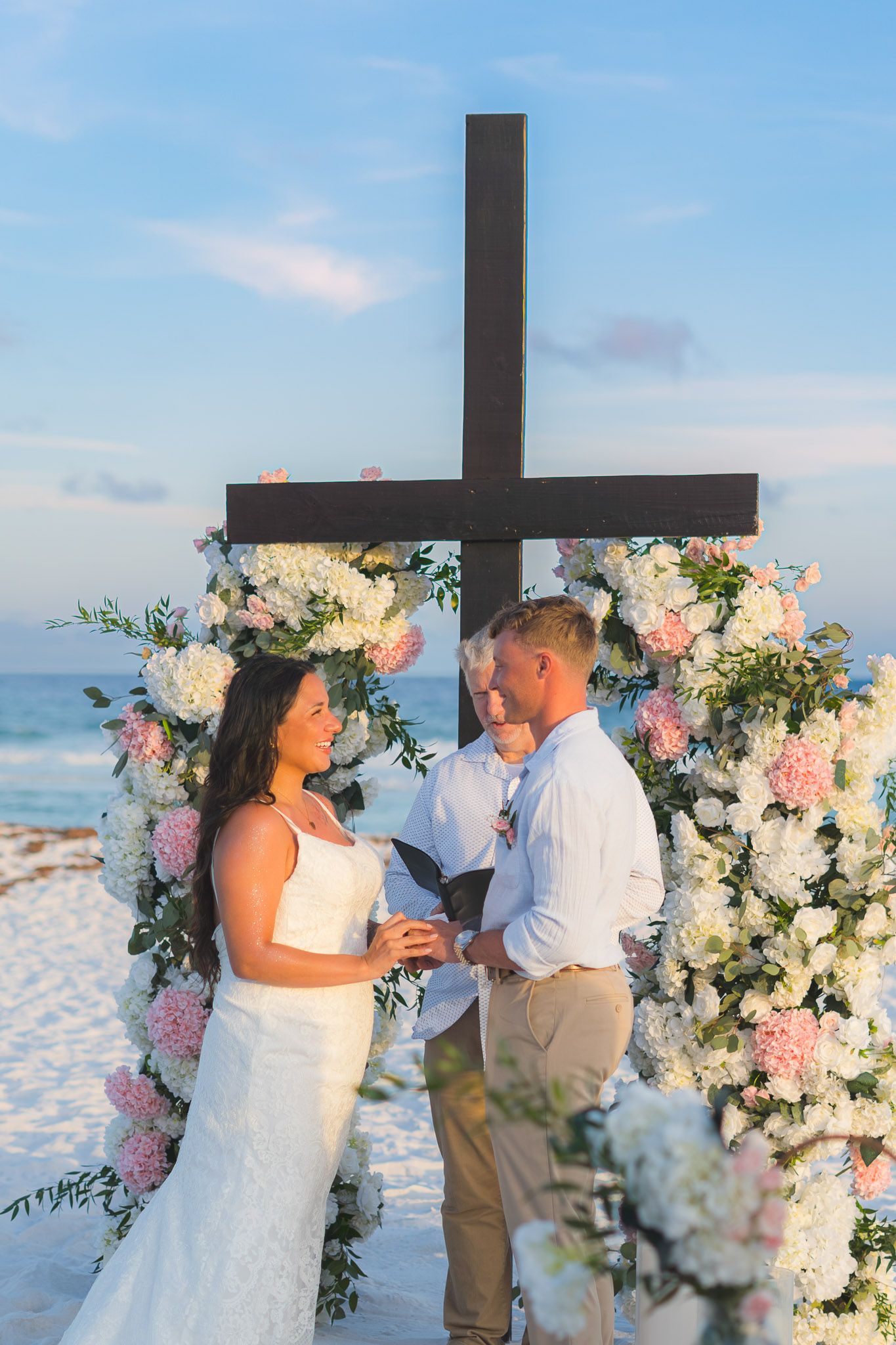 Couple exchanging vows at a beach wedding ceremony beneath a floral arch with a cross, ocean in the background.