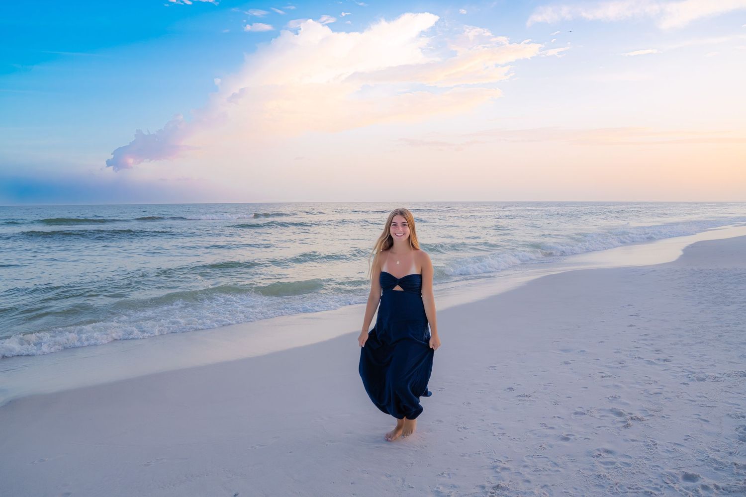 A blonde woman in a navy maxi dress walks barefoot on a sandy beach at sunset with gentle waves behind her.