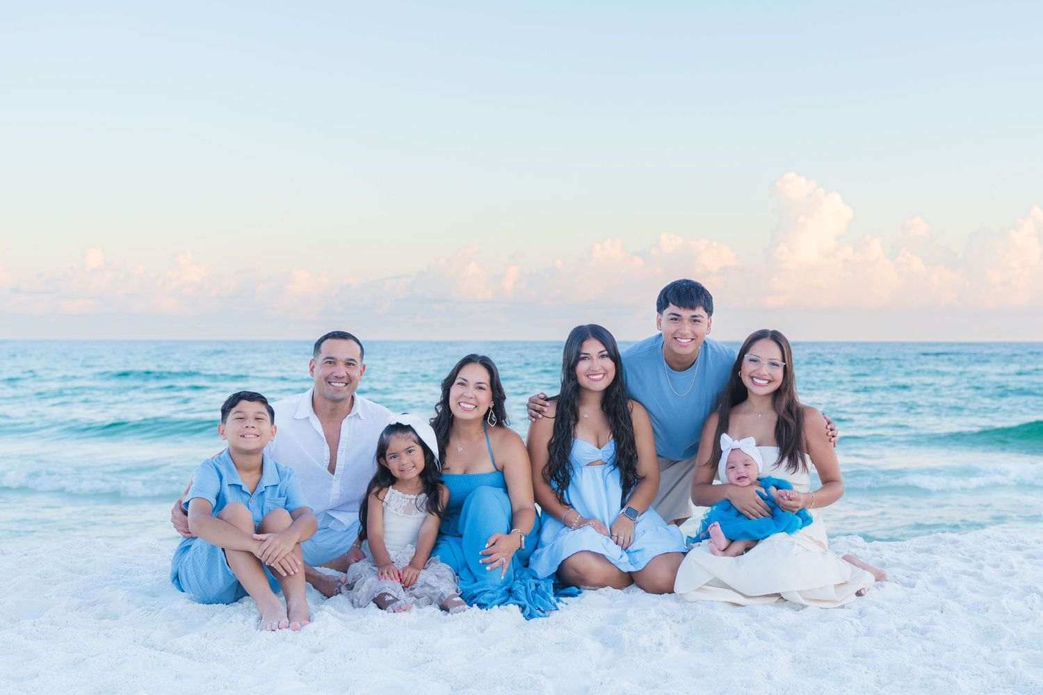 Family of seven posing together on a sandy beach at sunset, wearing coordinating blue and white outfits.
