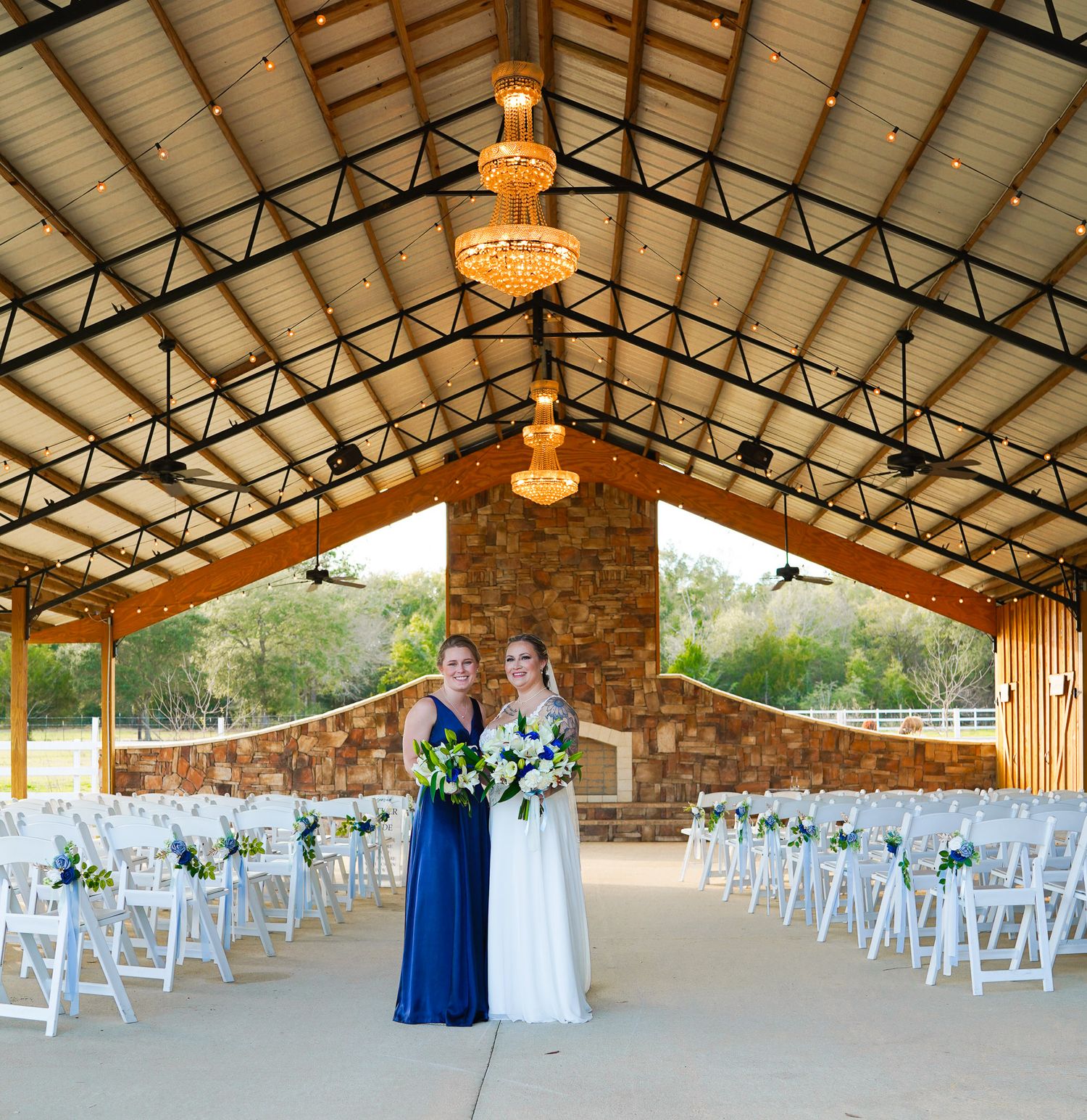 Bride and bridesmaid in navy dress stand in elegant open-air wedding venue with white chairs and chandelier lighting.