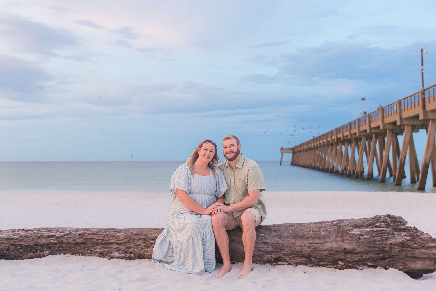 Smiling couple sitting on driftwood on a white sand beach with a wooden pier in the background.