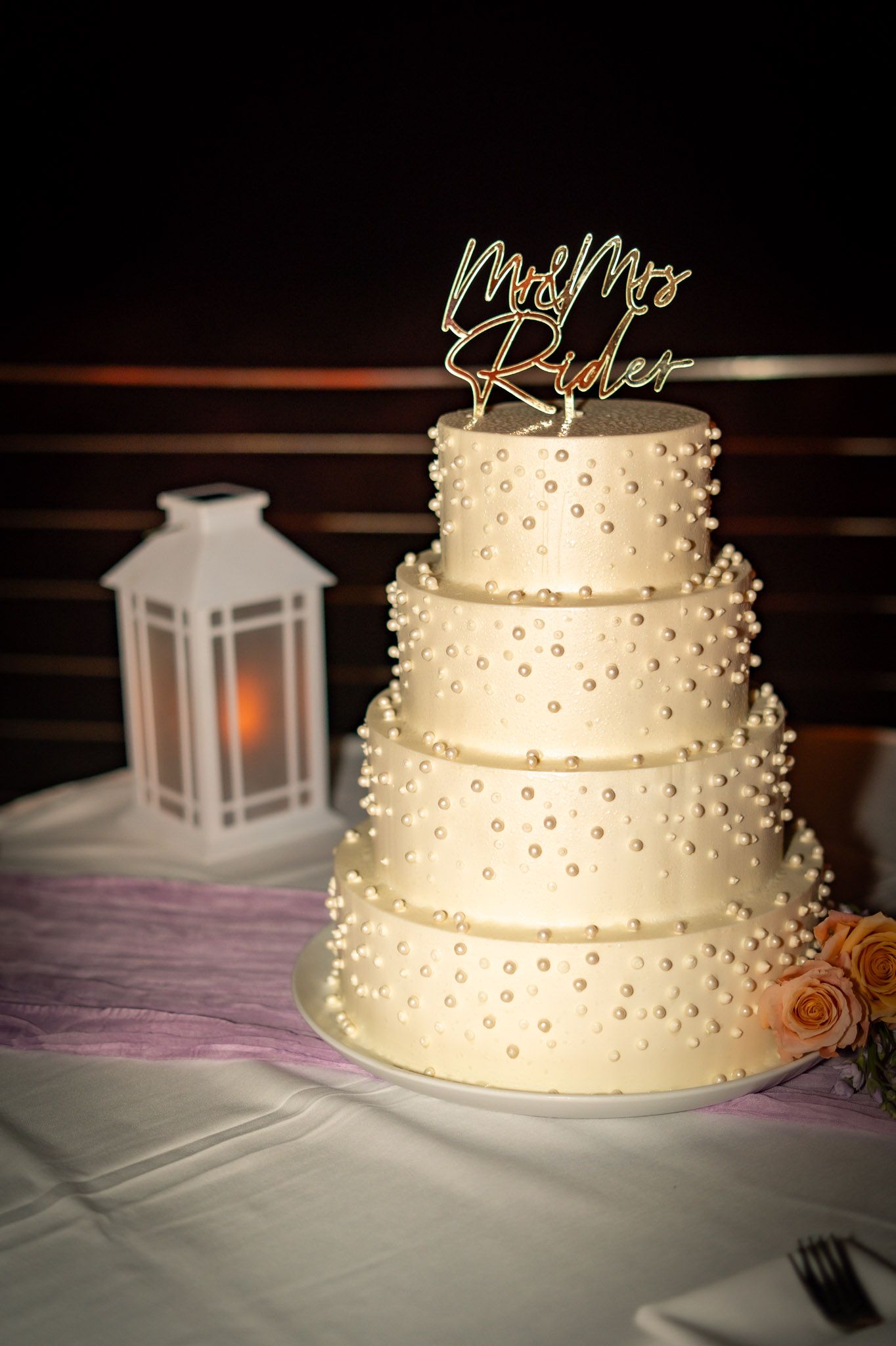 Four-tier white wedding cake with pearl details and Mr & Mrs Ryder gold topper, displayed beside a glowing lantern.