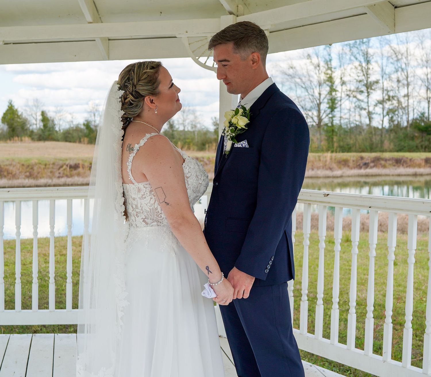 Bride and groom hold hands and gaze at each other in white gazebo with pond backdrop.