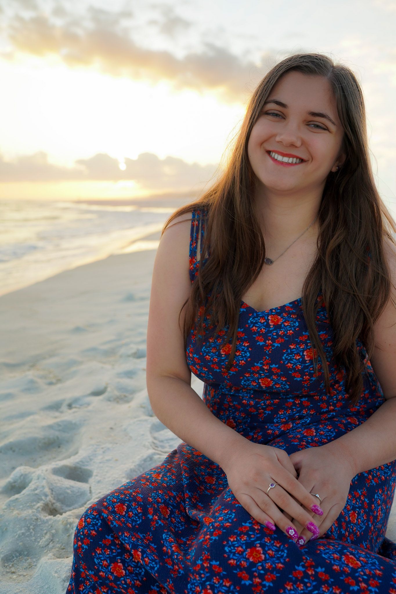 Smiling young woman in blue floral dress sitting on beach at golden sunset.