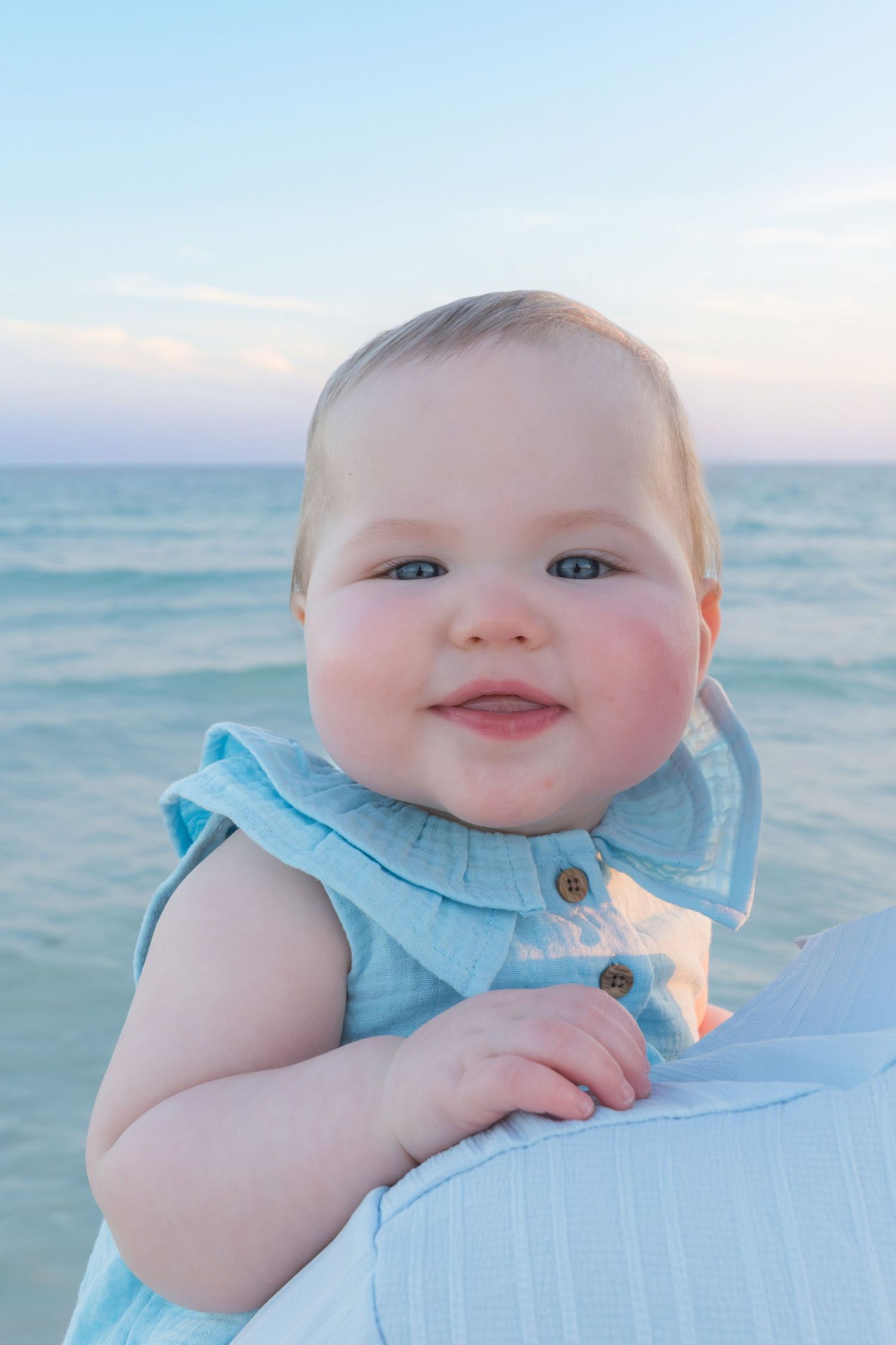 Smiling baby in light blue dress with blue eyes sitting by the ocean at sunset.