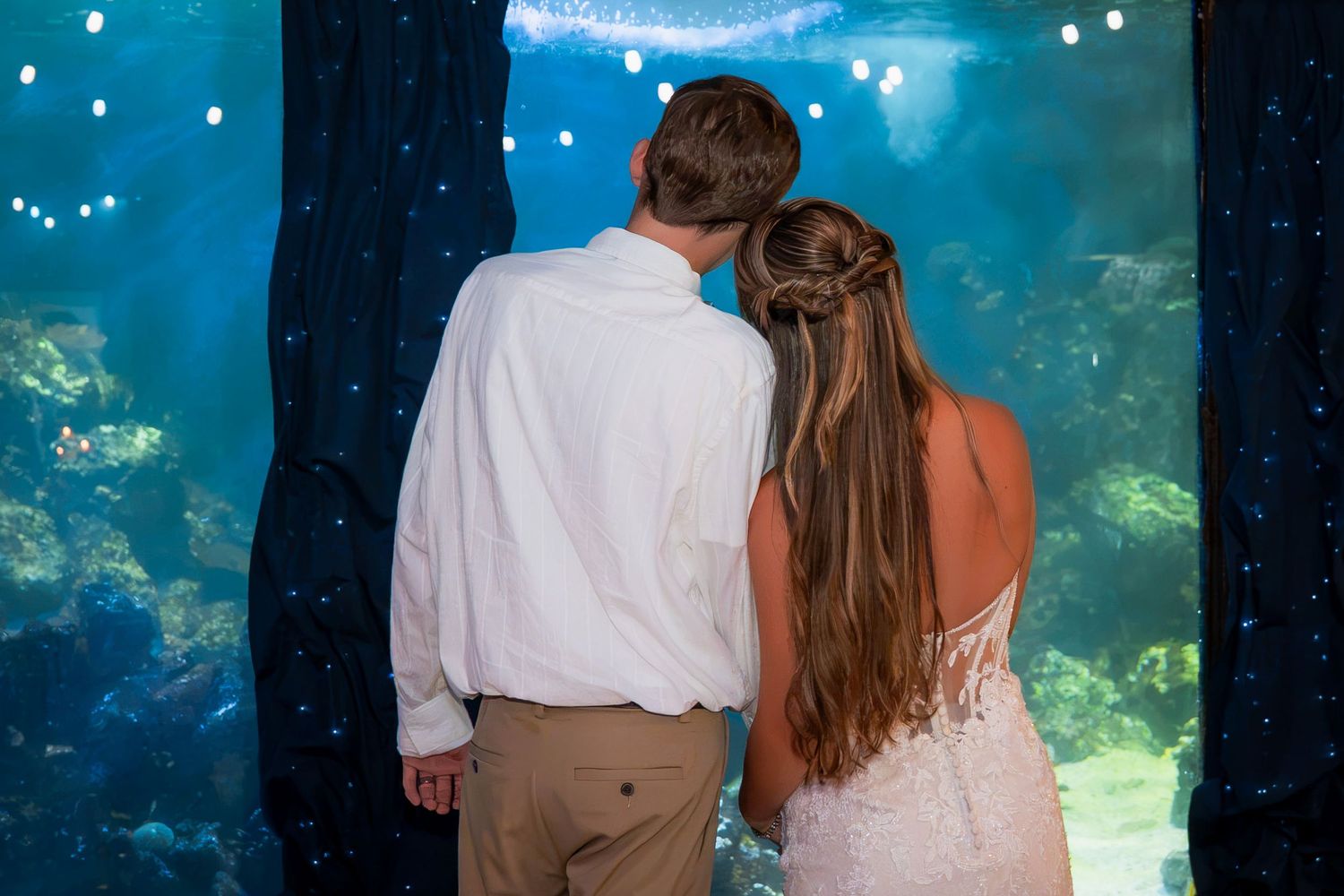 A bride and groom share a romantic moment gazing into a large illuminated aquarium tank together.