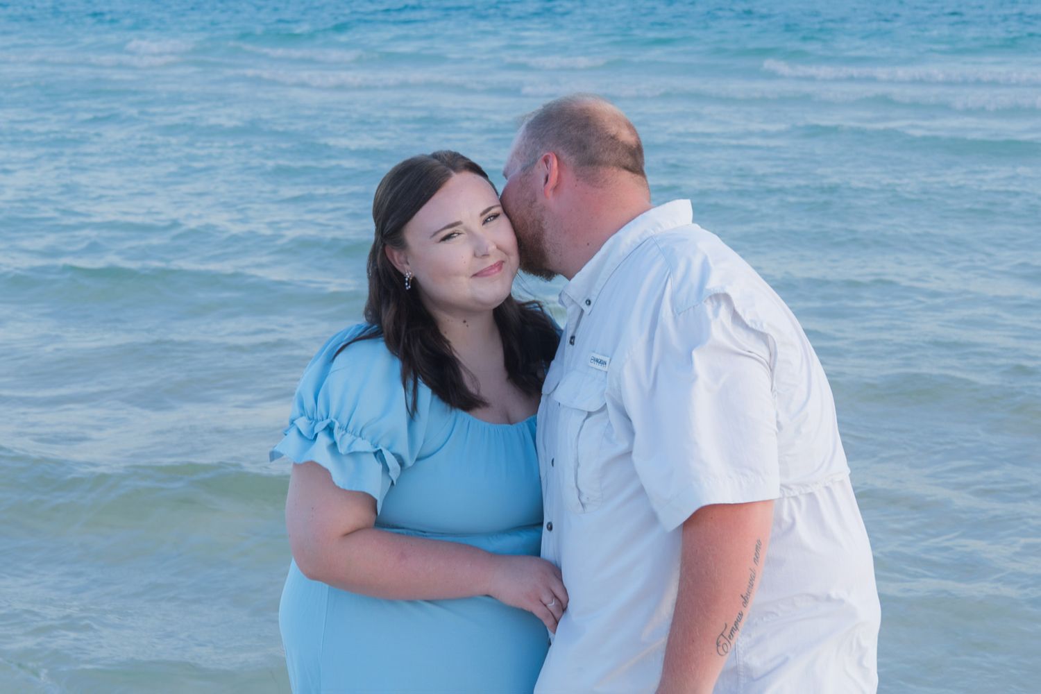 Pregnant couple embracing on a beach, woman in blue dress, man in white shirt, with ocean waves in the background.