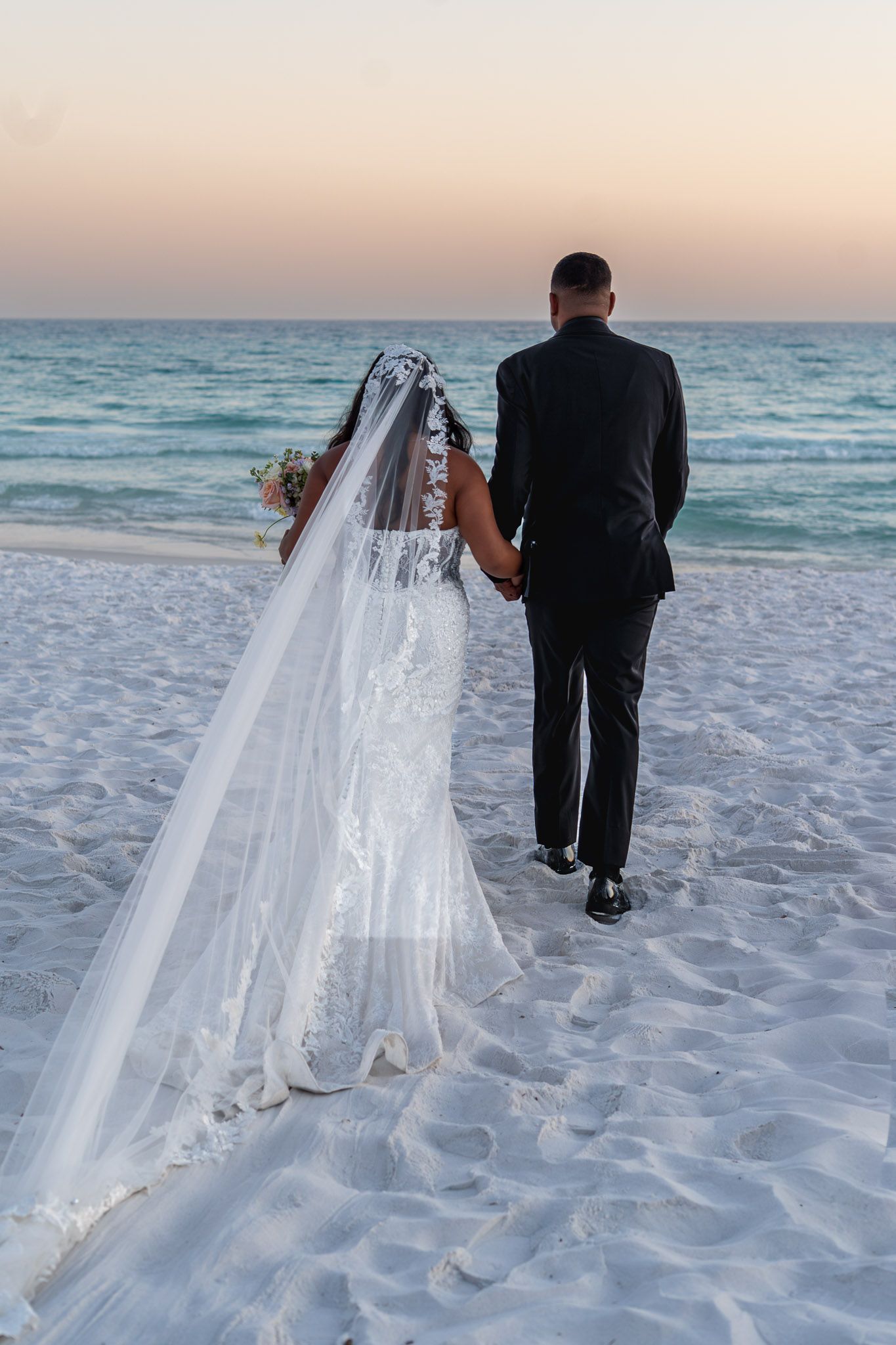 Bride and groom holding hands on white sand beach at sunset, bride wearing lace gown with long veil.