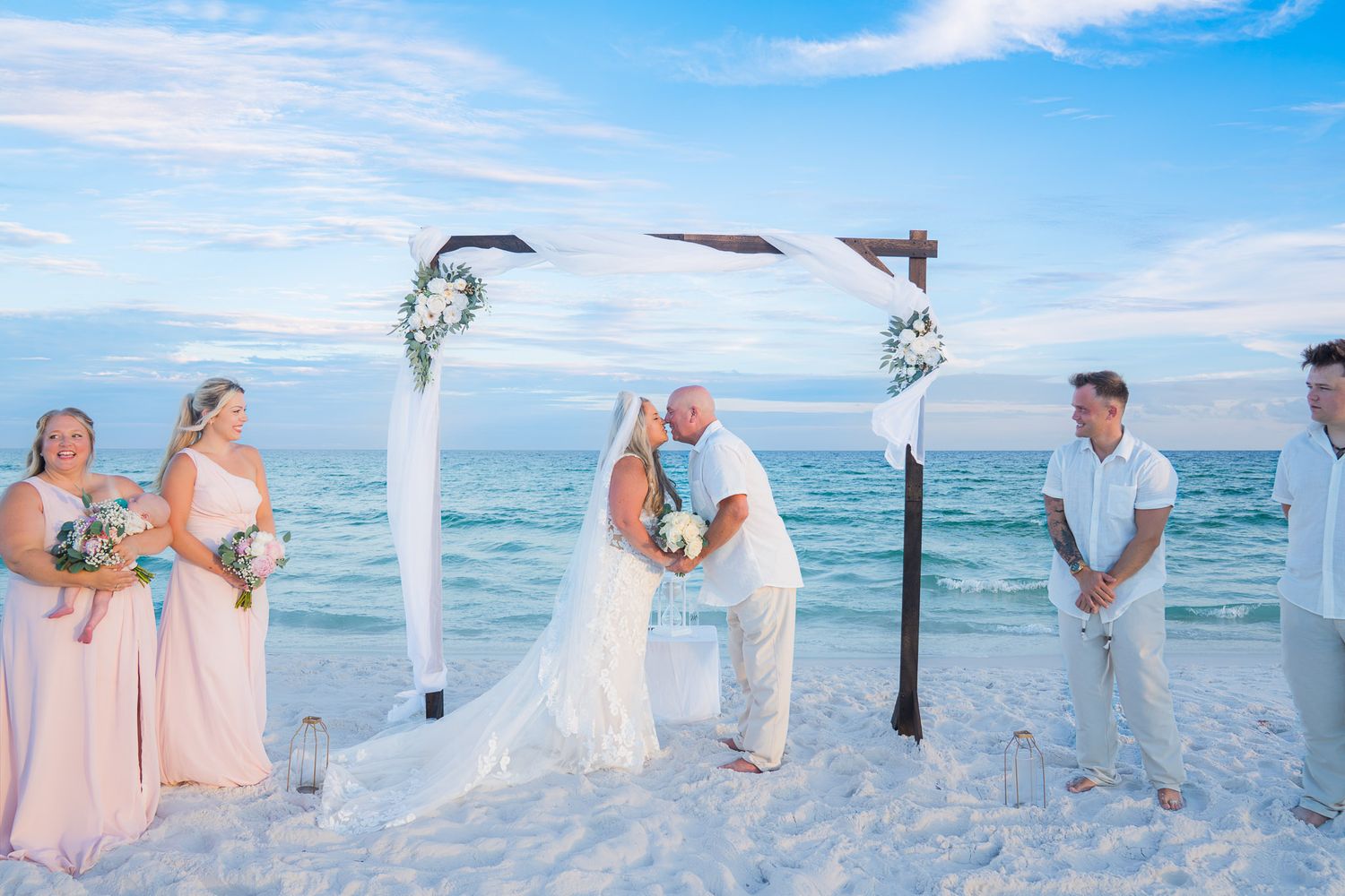 Couple sharing first kiss at beach wedding ceremony with bridesmaids and groomsmen under floral arch on white sand.