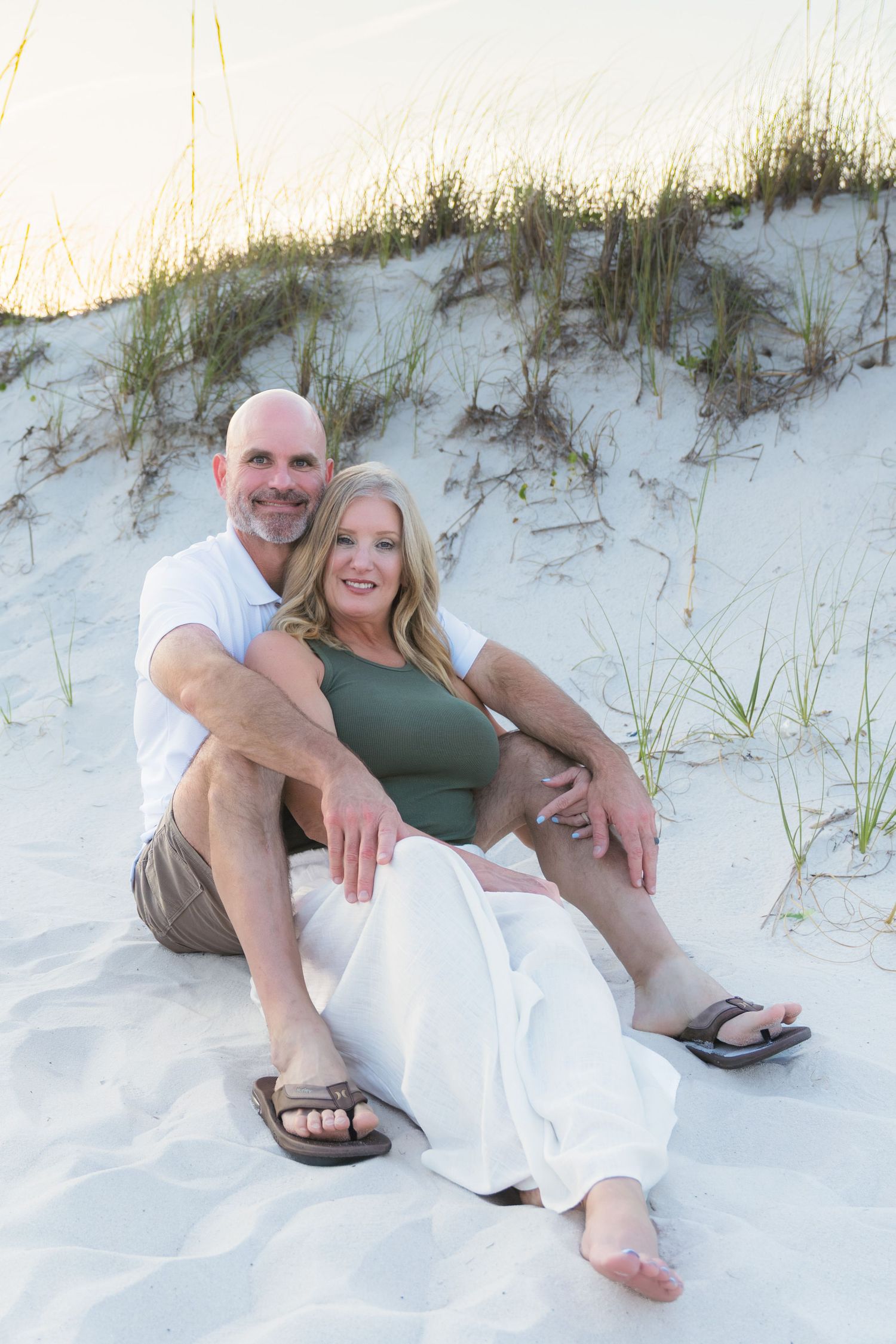 Couple sitting on white sand dunes at beach, woman in green top and white skirt, man in white shirt and khaki shorts.