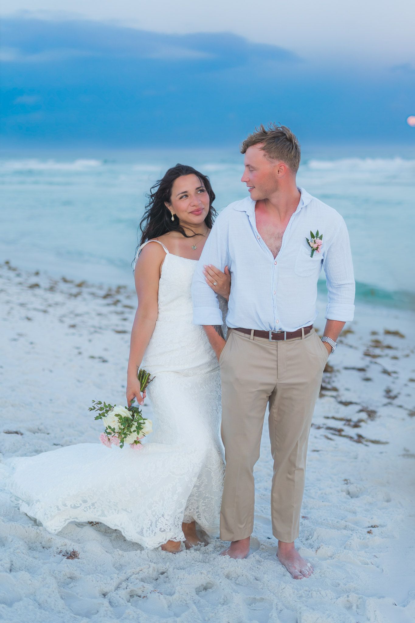 Bride and groom standing together on a white sand beach at dusk.