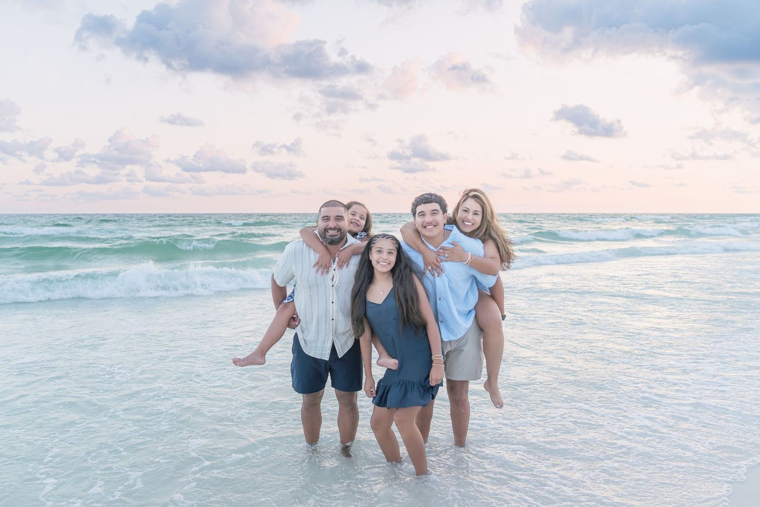 Family of five smiling together on a sandy beach at sunset with calm turquoise ocean waves.