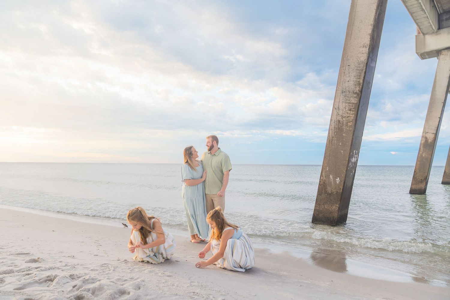 Family with two young girls playing in sand near beach pier at dusk.