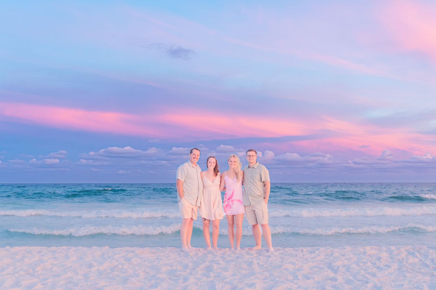 Family of four posing on a white sand beach at sunset with vibrant pink and purple sky over turquoise ocean.