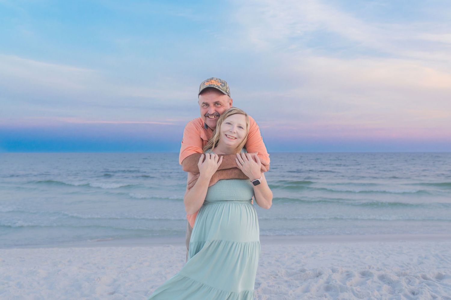 Pregnant woman in mint dress embraced by partner on a serene beach at sunset.